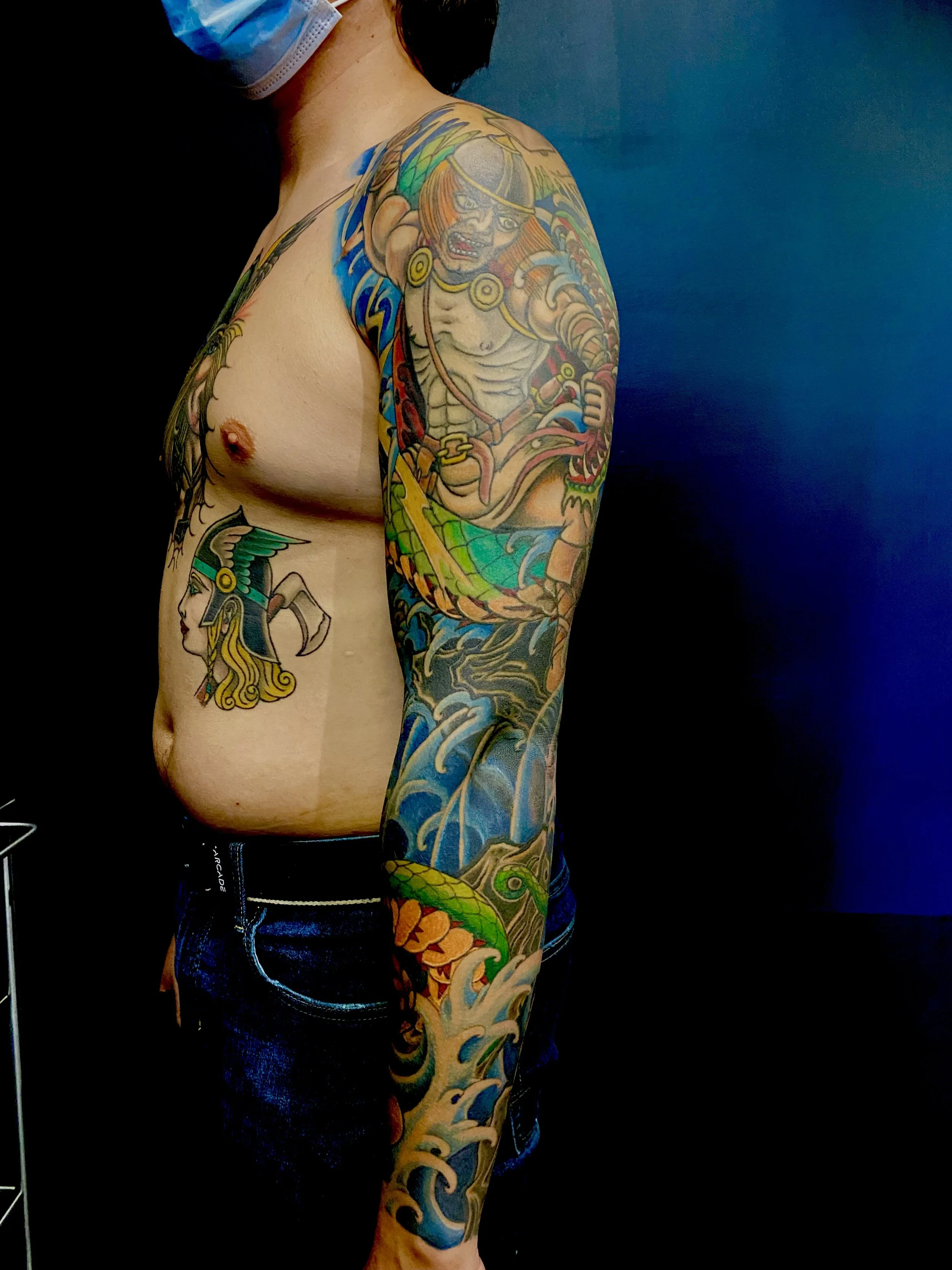 A person with tattoos on their arm and torso, wearing a face mask and dark clothing, standing against a dark background.