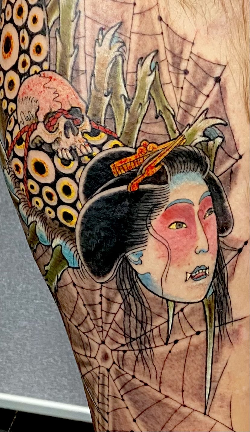 Tattoo of a geisha with elaborate hair decorations, a pale face, and vivid makeup, accompanied by a skull and spider webs in the background.