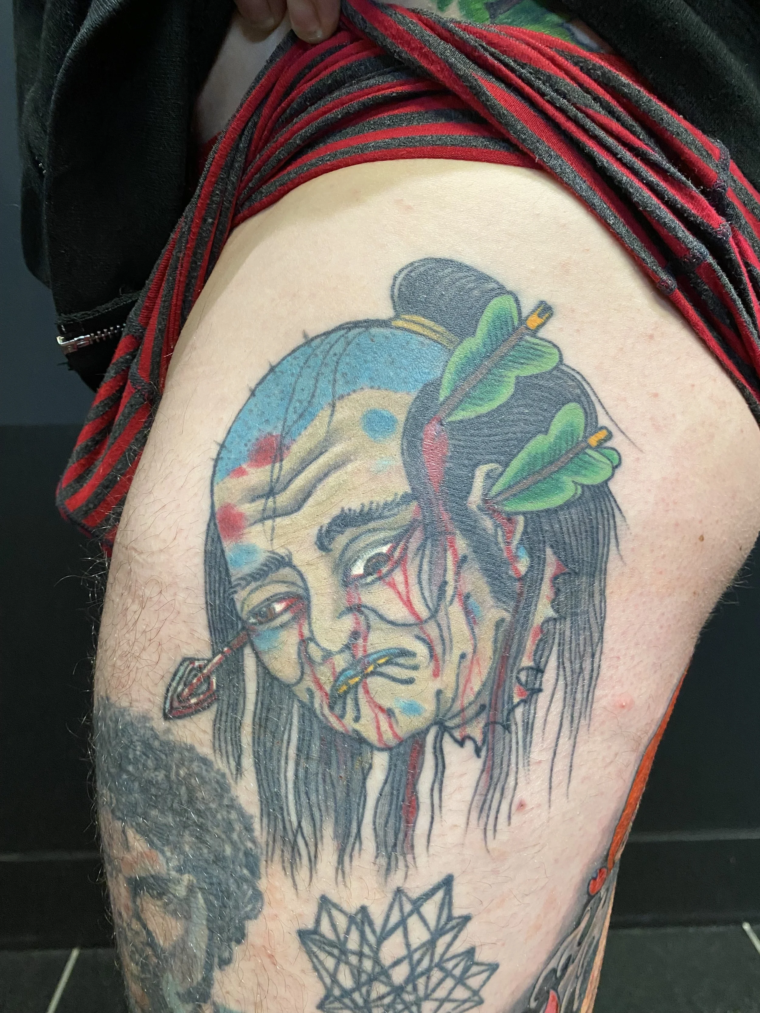 Tattoo of a Native American woman's face with an arrow in her head, accompanied by a green leaf and red accents, on a person's upper arm.
