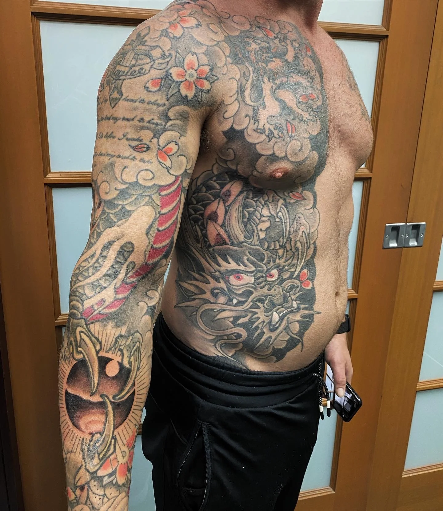 A shirtless man with extensive tattoos on his chest, arm, and torso, standing in a room with wooden doors and panels.