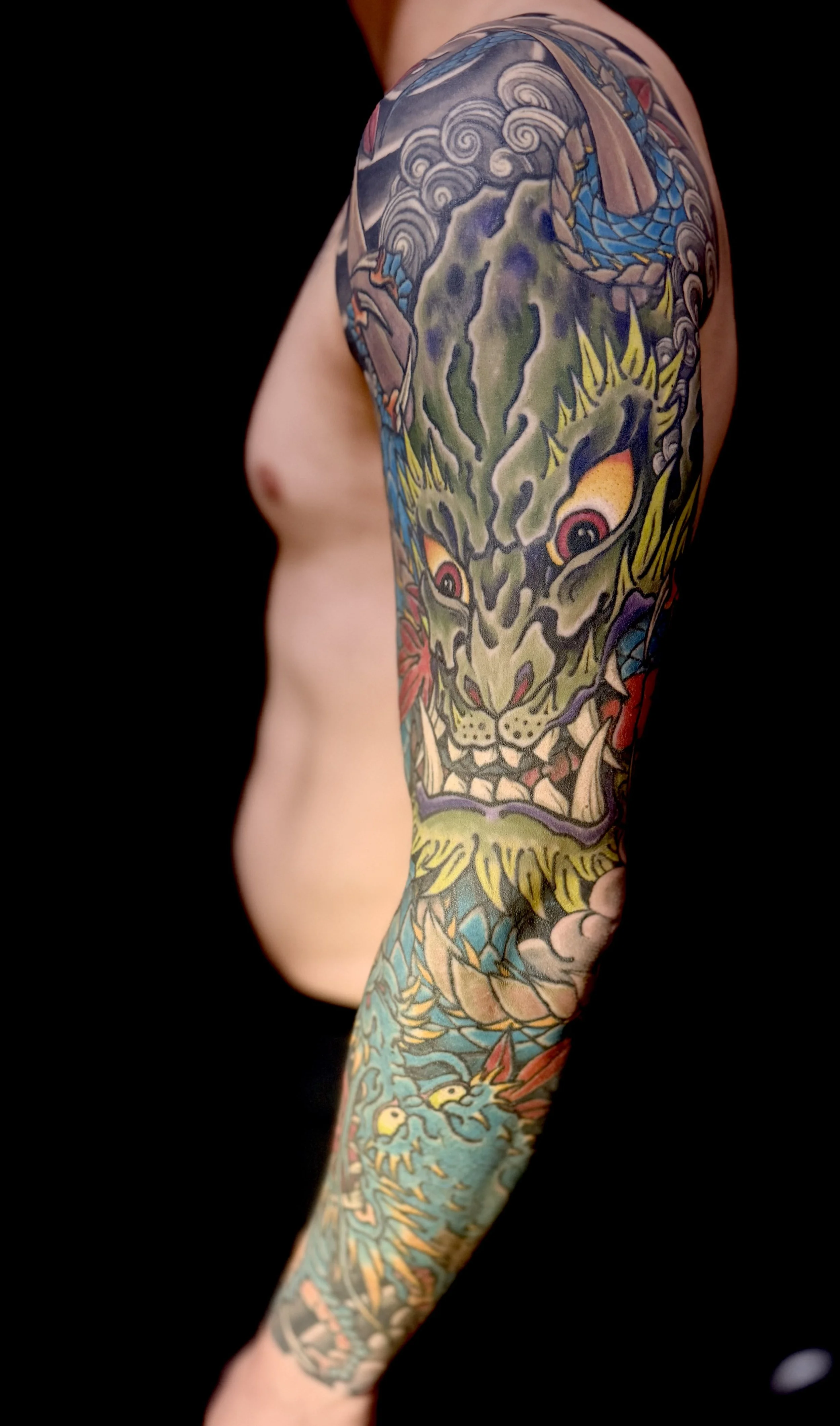 Close-up of a person's arm with a colorful Japanese-style tattoo featuring a fierce dragon face with sharp teeth, yellow eyes, and swirling clouds on a black background.