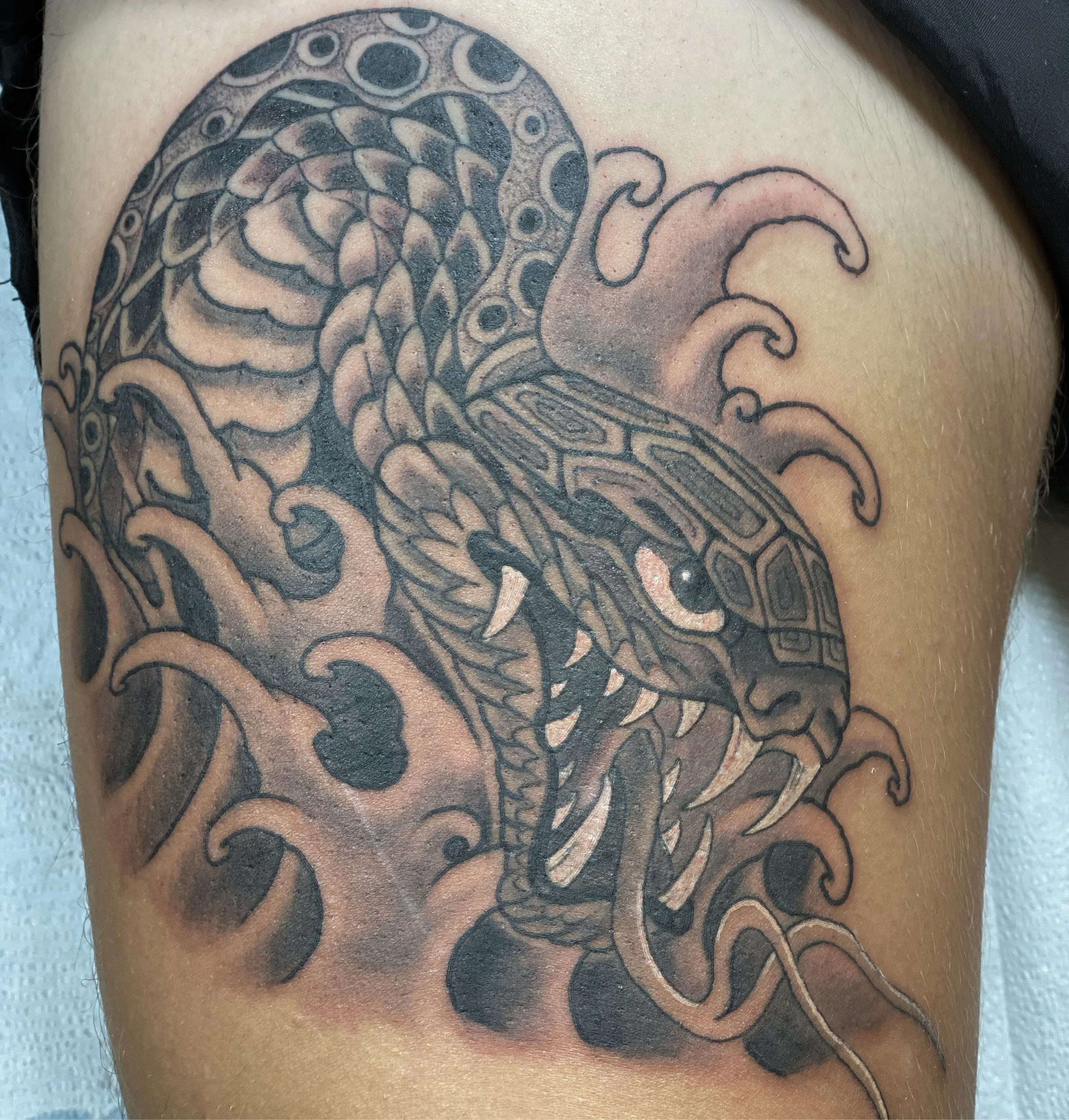 Black and gray tattoo of a snake with detailed scales, surrounded by stylized waves on a person's arm.
