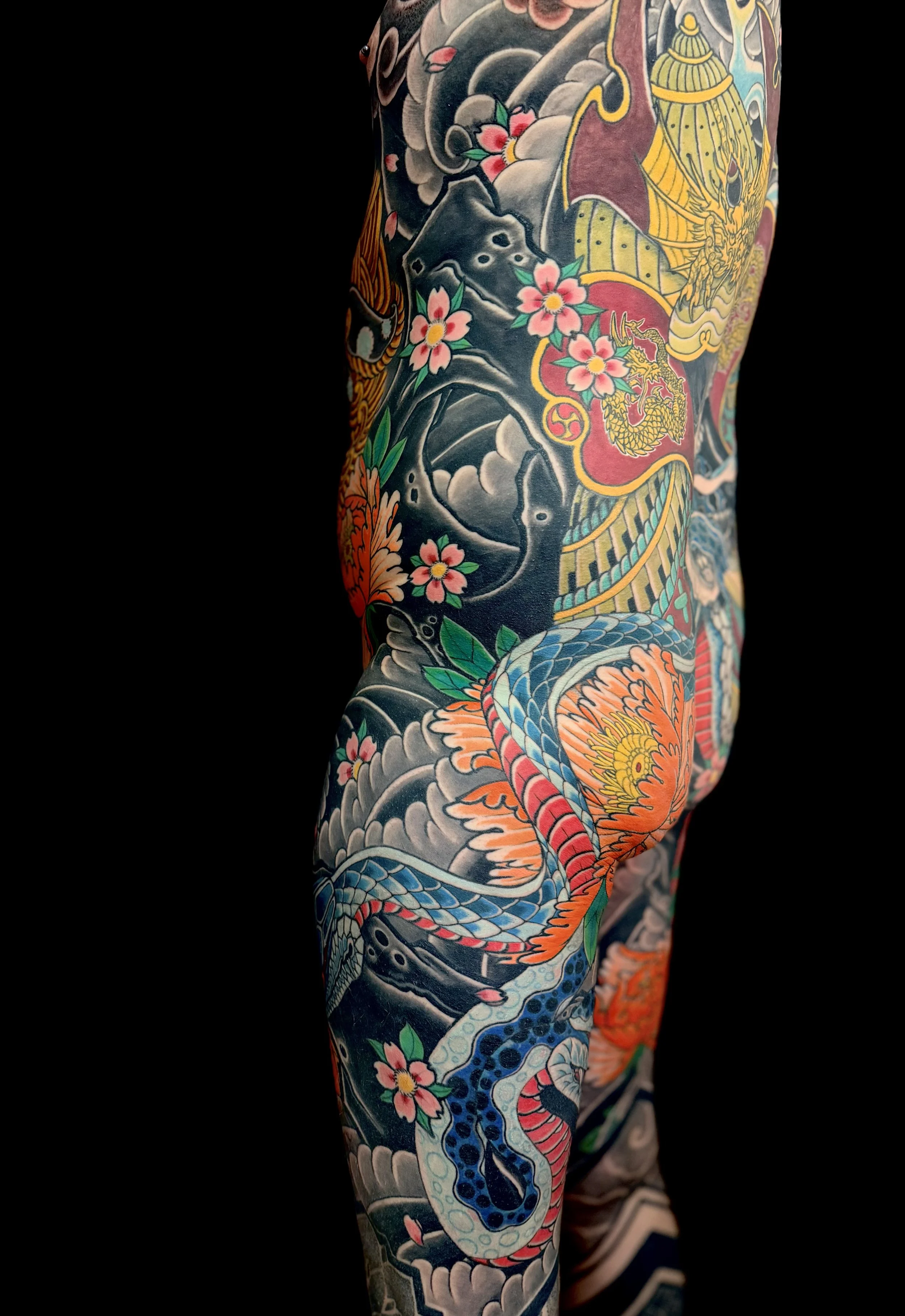 Close-up of a person's arm covered in a colorful traditional Japanese-style tattoo, featuring a dragon, cherry blossoms, clouds, and waves, against a black background.