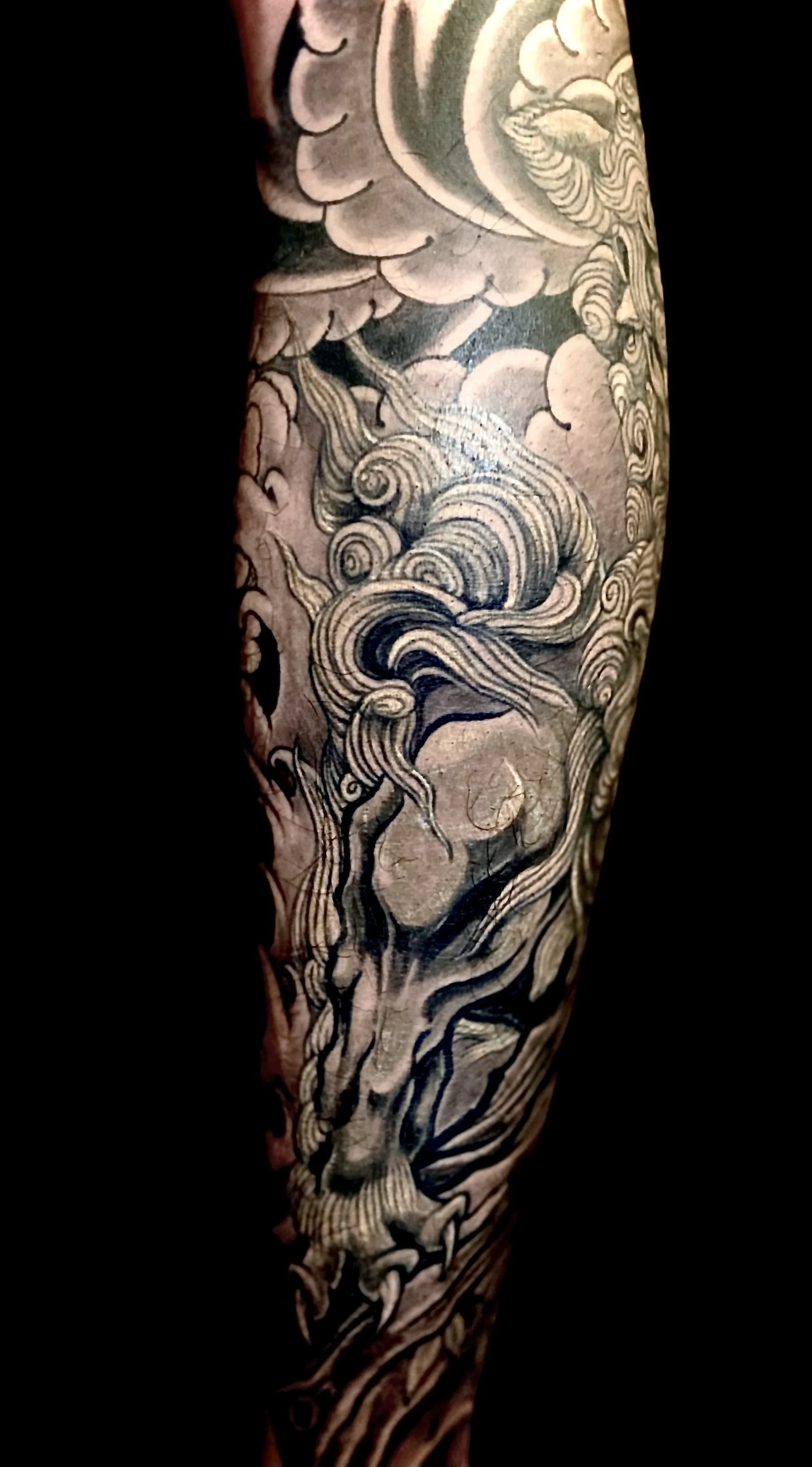 A detailed black and gray tattoo of a mythological or fantasy creature with flowing mane, clouds, and swirling elements on a person's arm.
