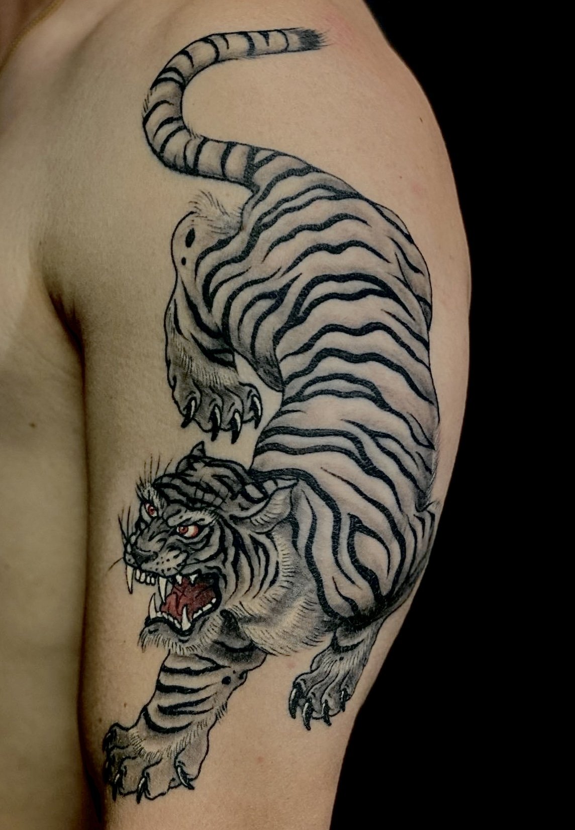 Tattoo of a tiger in black and gray with red eyes, showing a roaring face and a full body, inked on a person's upper arm.