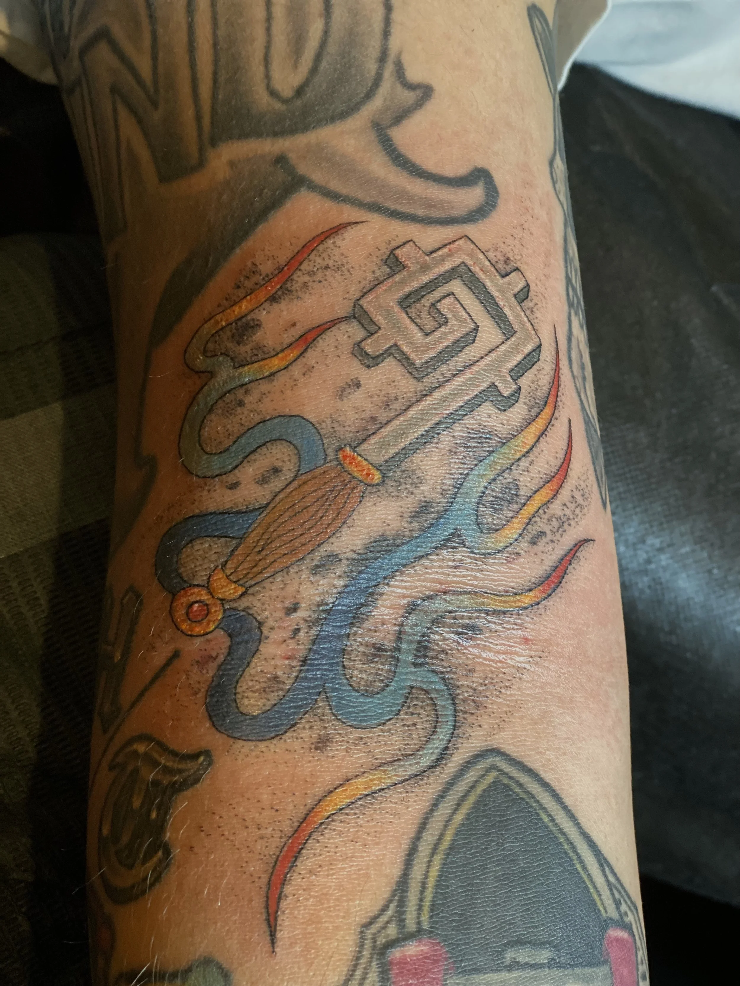 Tattoo of a stylized sword with a spiral design on the blade, flames in orange, yellow, and red colors, and a wooden handle, on a person's arm.