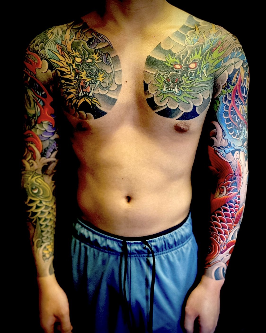 Tattooed man with colorful dragon tattoos on both arms and chest, wearing blue shorts.