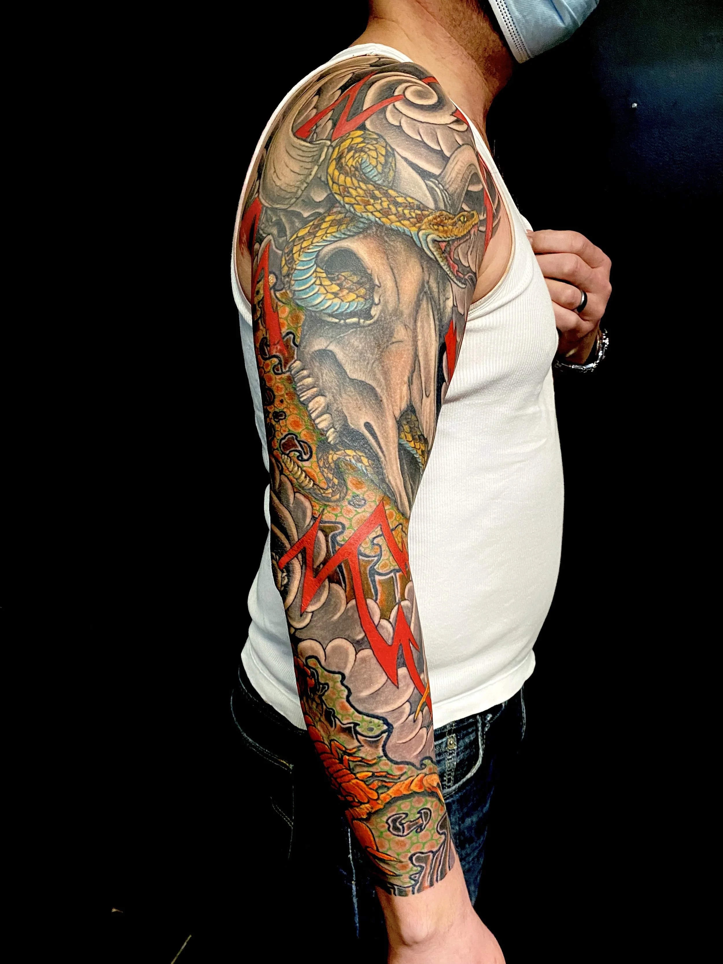 A person with a colorful sleeve tattoo featuring a snake, a skull, and cloud-like patterns, wearing a white tank top and blue jeans, against a black background.
