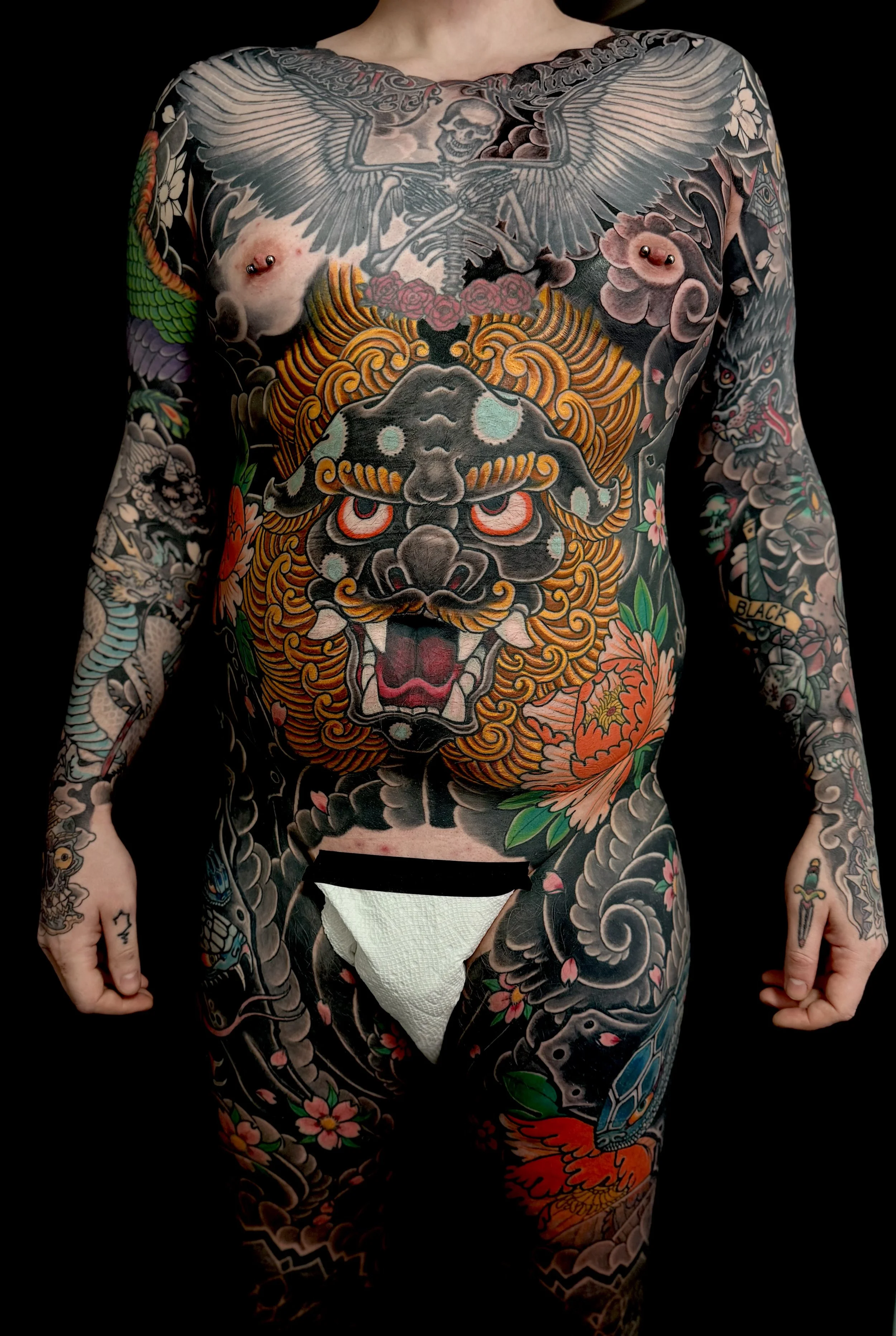 Tattoos covering a person's chest, arms, and legs, including a large wolf-head, a skull with wings, a tiger face, flowers, dragons, and other intricate designs.