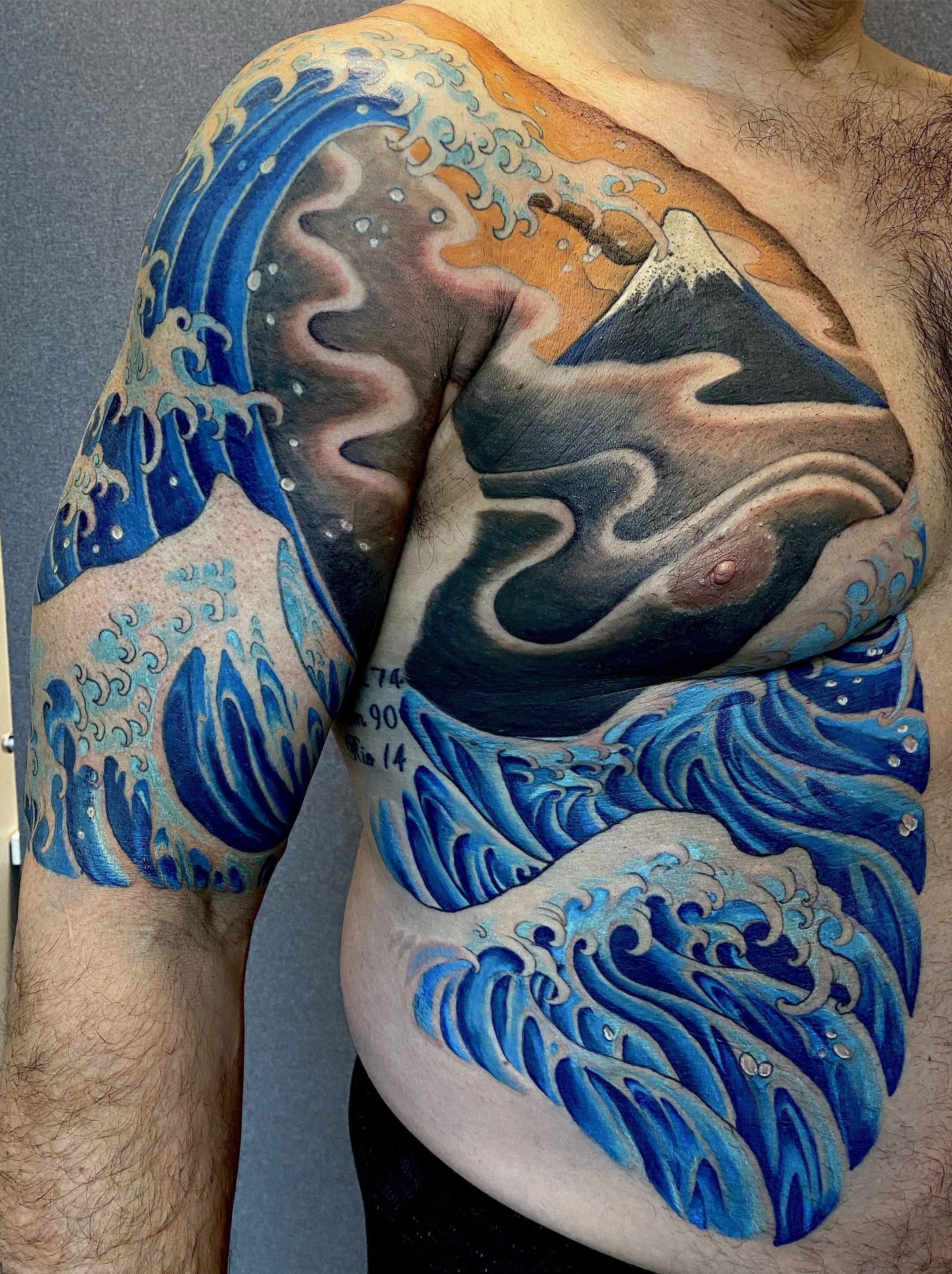 Colorful tattoo of a large ocean wave and mountain, inspired by Hokusai's "The Great Wave off Kanagawa," on a man's shoulder and upper arm.