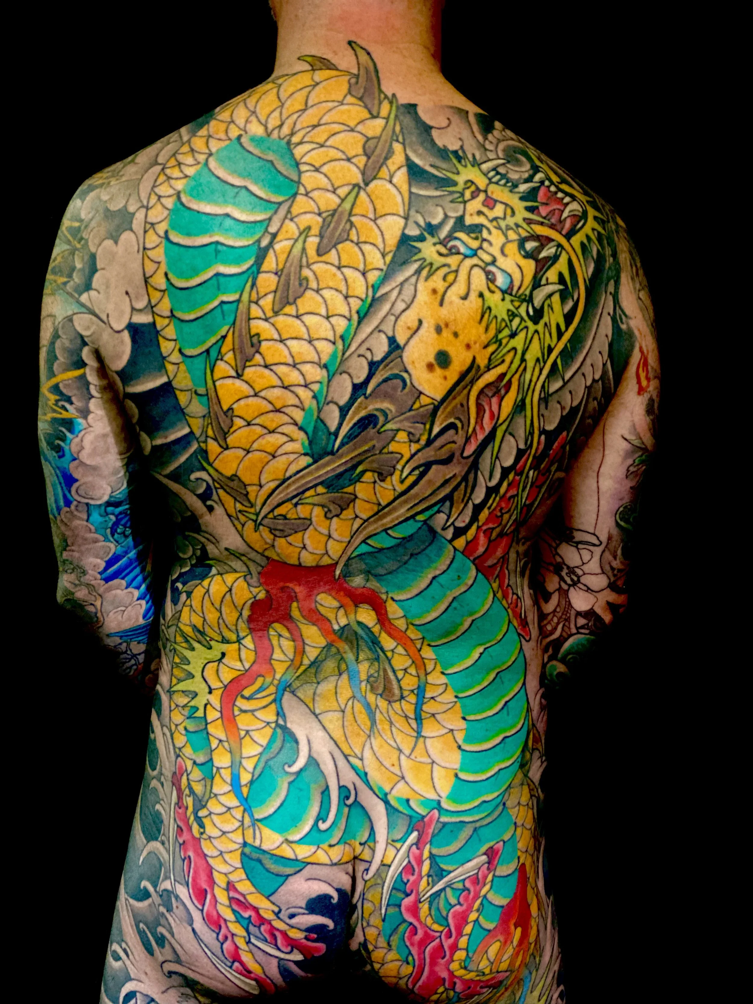 Back view of a person with extensive Japanese-style tattoo art covering the entire back, depicting a large yellow and green dragon intertwined with clouds, flames, and water elements.