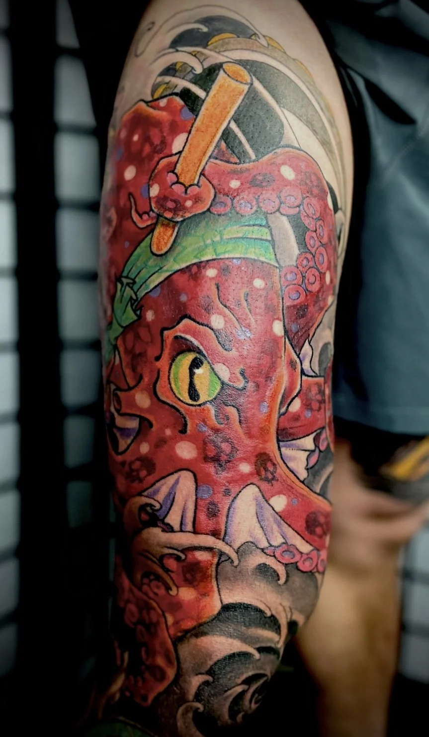 Colorful tattoo of a red octopus with yellow eyes holding a stick, covering most of the upper arm, with waves and clouds in the background.