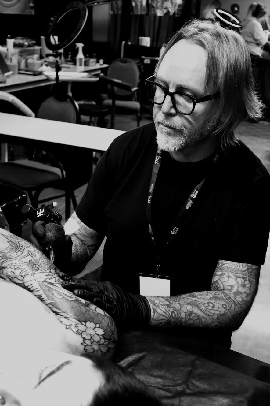 A man with glasses and shoulder-length hair tattooing a person's arm in a tattoo studio, with tattoo equipment and chairs in the background.