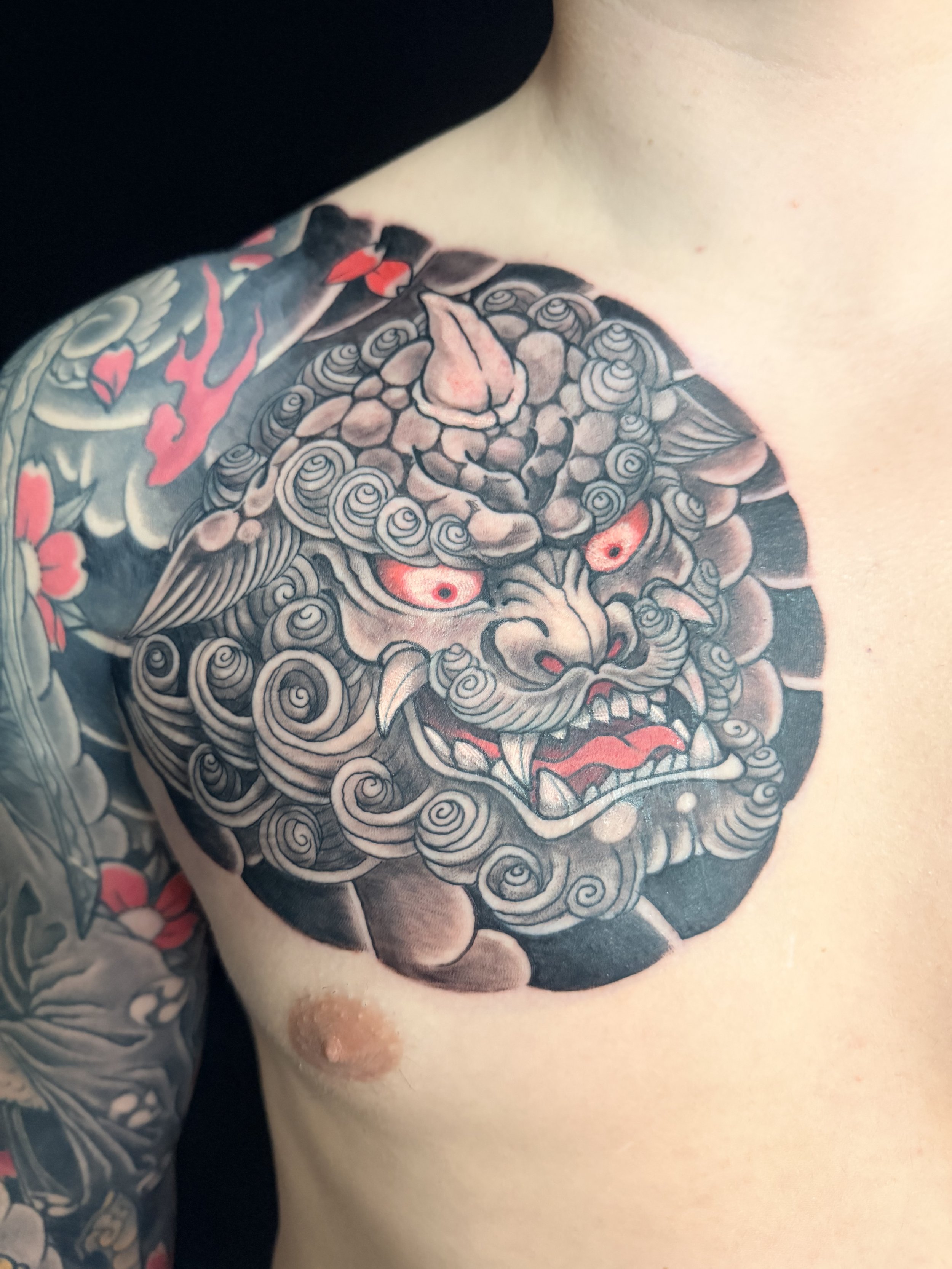 Close-up of a traditional Japanese tattoo of a fierce face with swirling patterns, red eyes, and elements of red and gray on a person's shoulder.