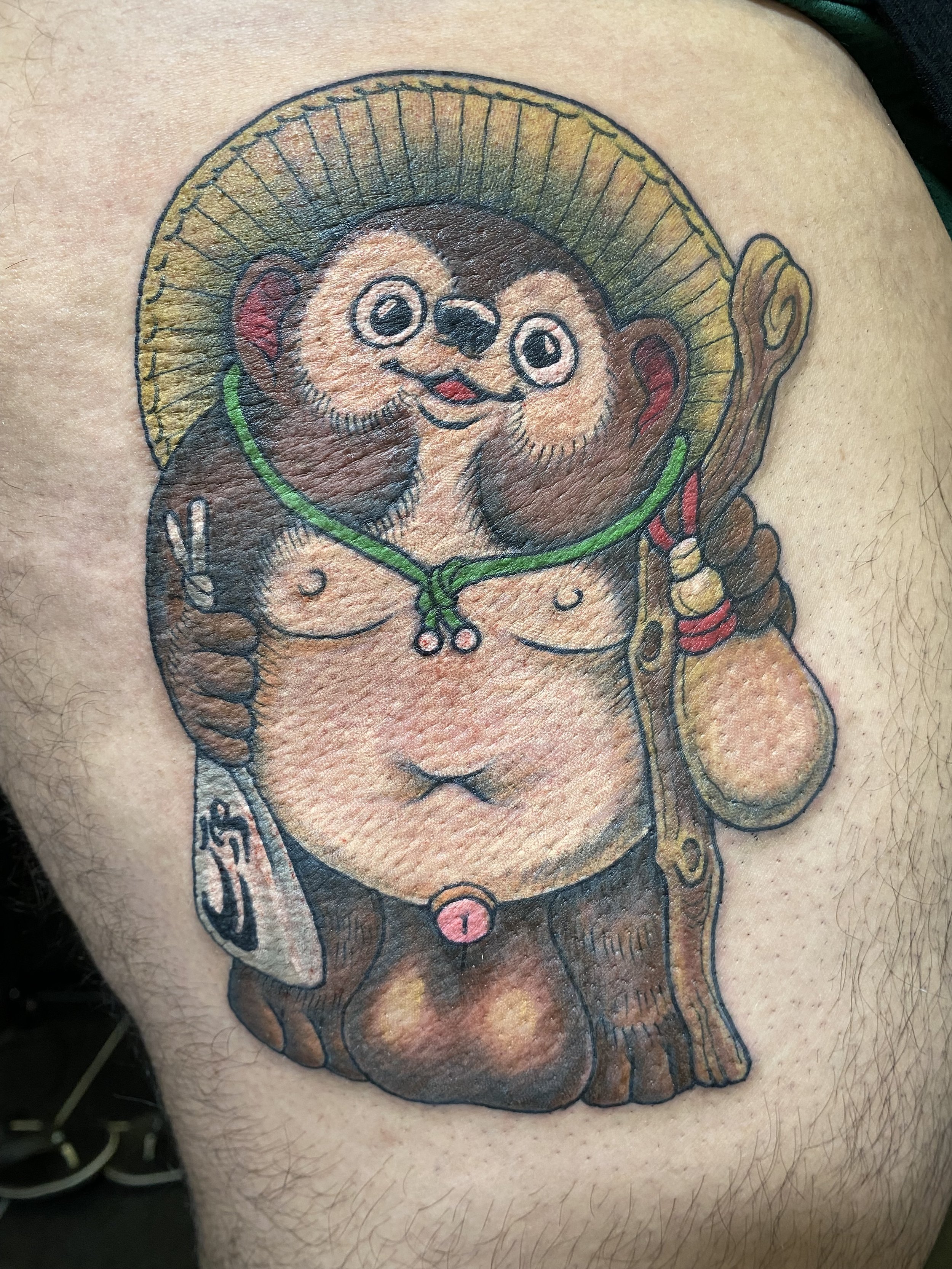 Colorful tattoo of a cartoon-style Buddha bear with a straw hat, holding a staff and a bag, with a happy face, on the skin of a person's upper arm.