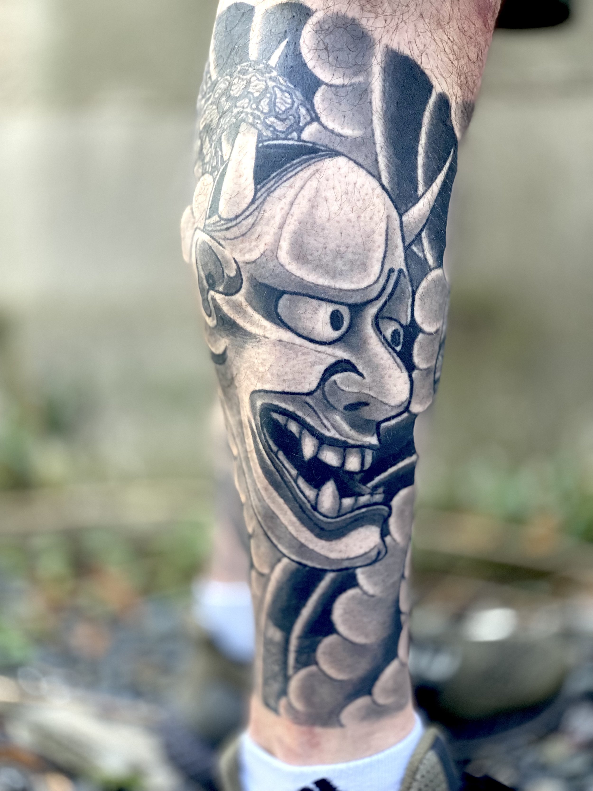 Black and gray tattoo of a fierce, angry face with sharp teeth, flowing hair, and exaggerated features on a person's leg.
