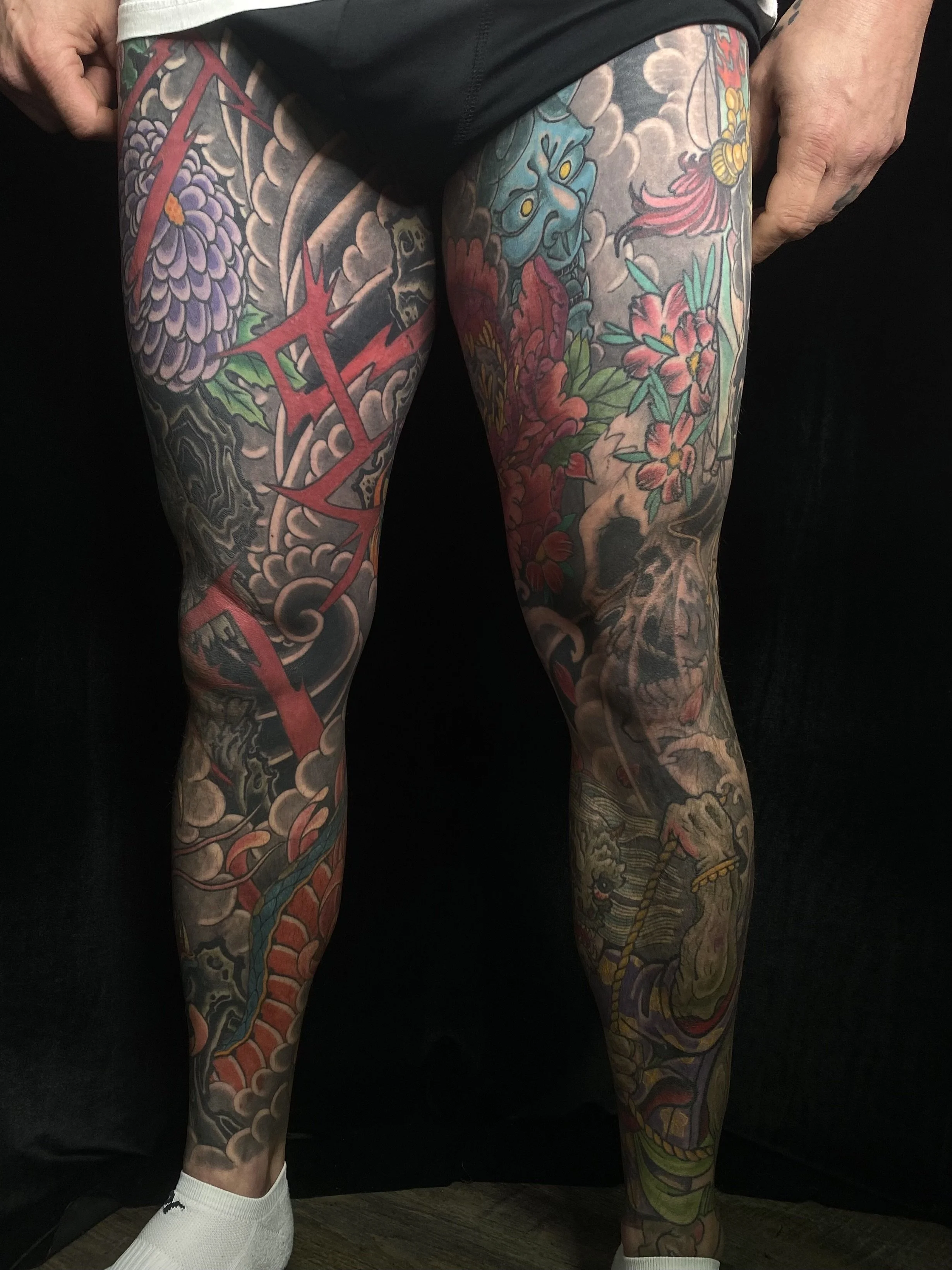 Colorful full-leg tattoos featuring various Japanese-style dragons, flowers, a blue oni mask, and a skull, covering both legs from thighs to ankles