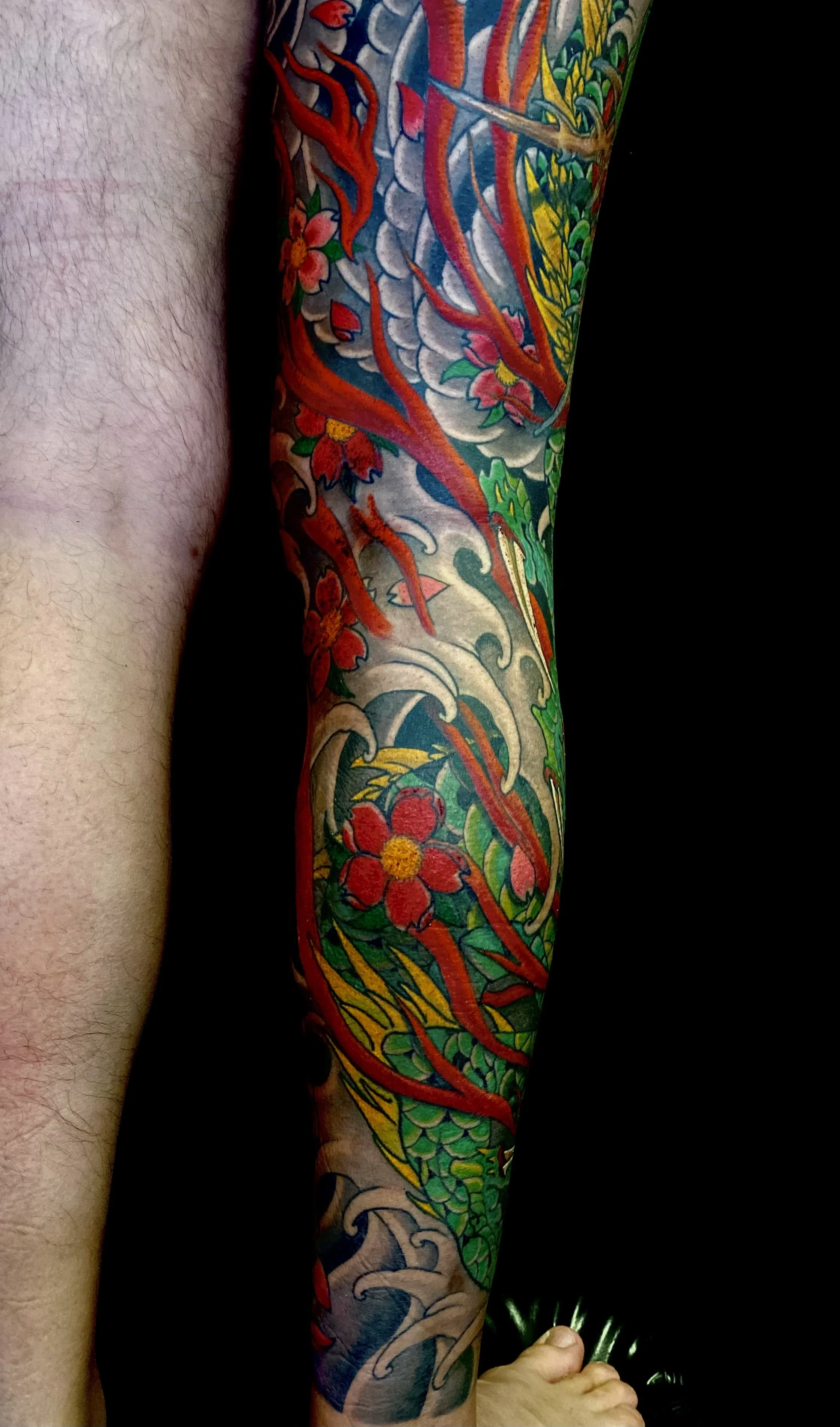A person's arm with a colorful Japanese-style tattoo featuring a dragon, flowers, and waves.