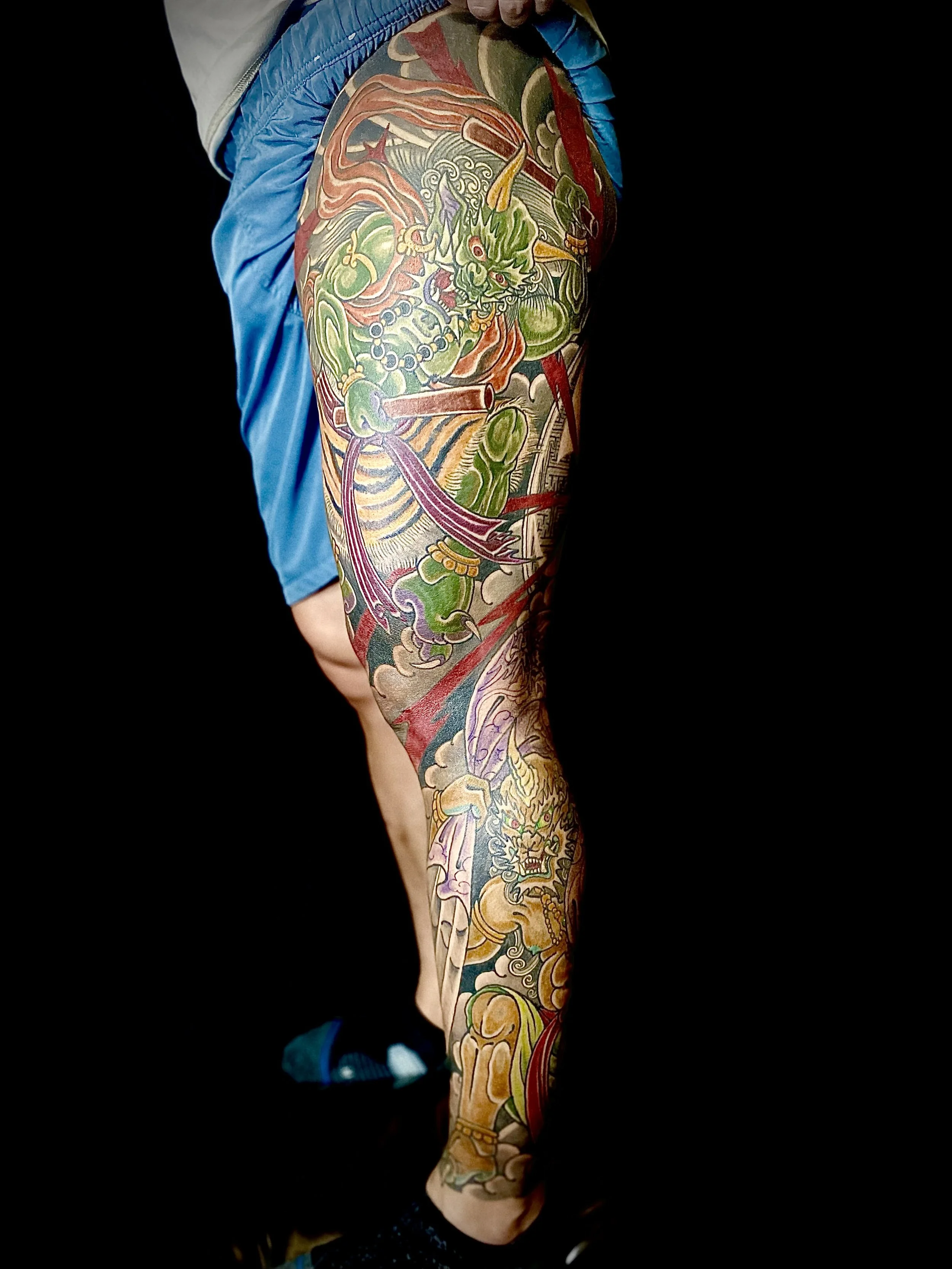 Tattooed leg with a colorful Japanese-style dragon and warrior design against a black background.