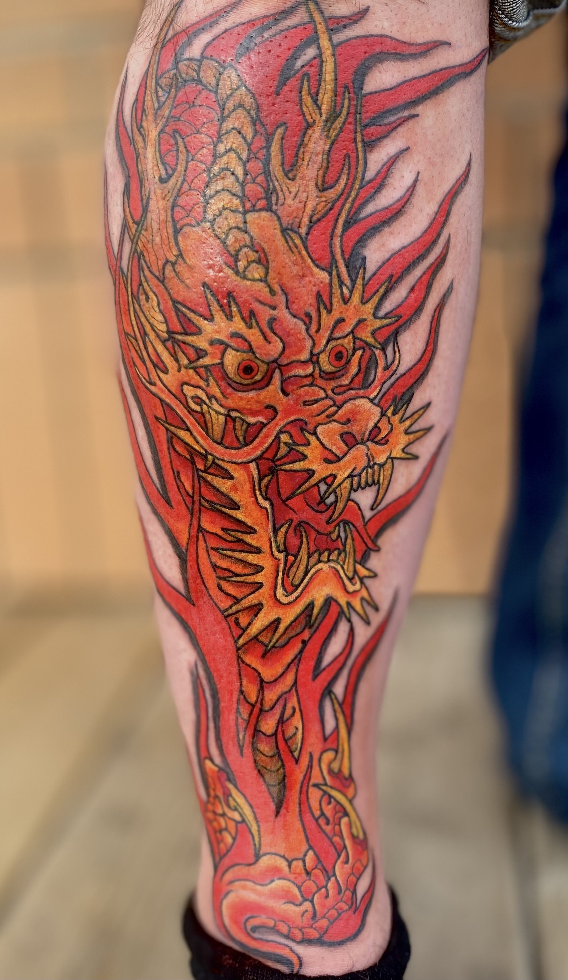 Tattoo of a fierce, fiery dragon in red, orange, and black ink covering the person's calf.