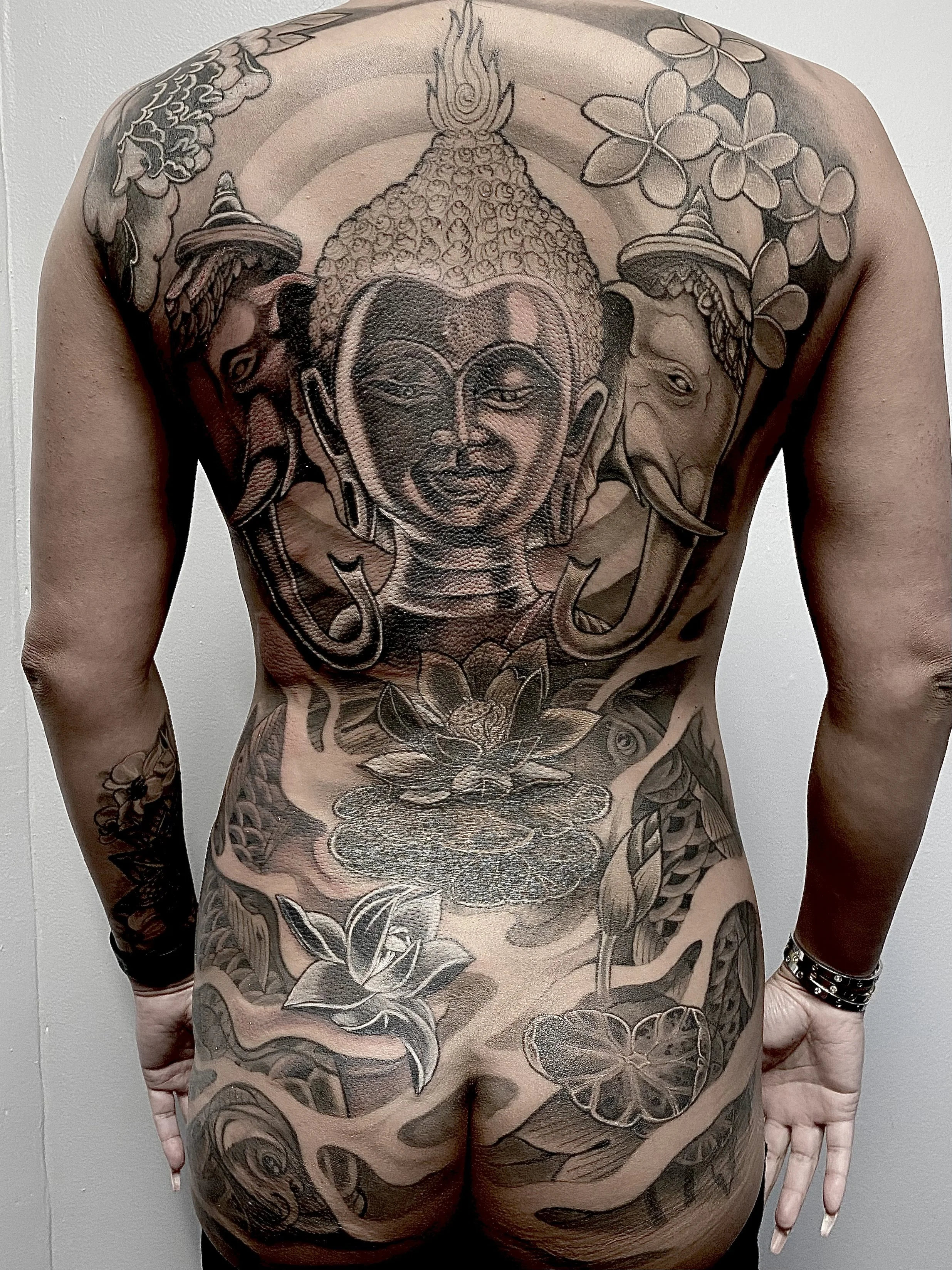 Back and upper body tattoos of a person featuring a large Buddha face, elephants, lotus flowers, and floral patterns in black and gray ink.