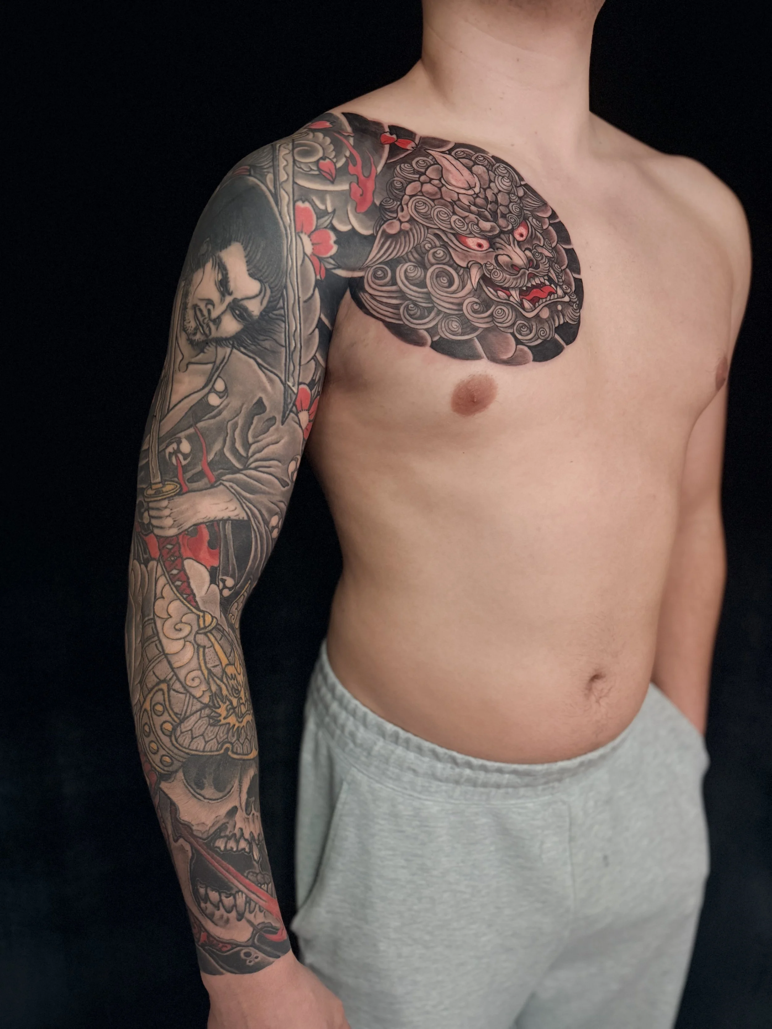 A person's upper arm and shoulder with detailed traditional Japanese style tattoos, including a fierce lion's face with red eyes and open mouth, surrounded by wave, cloud, and floral motifs.