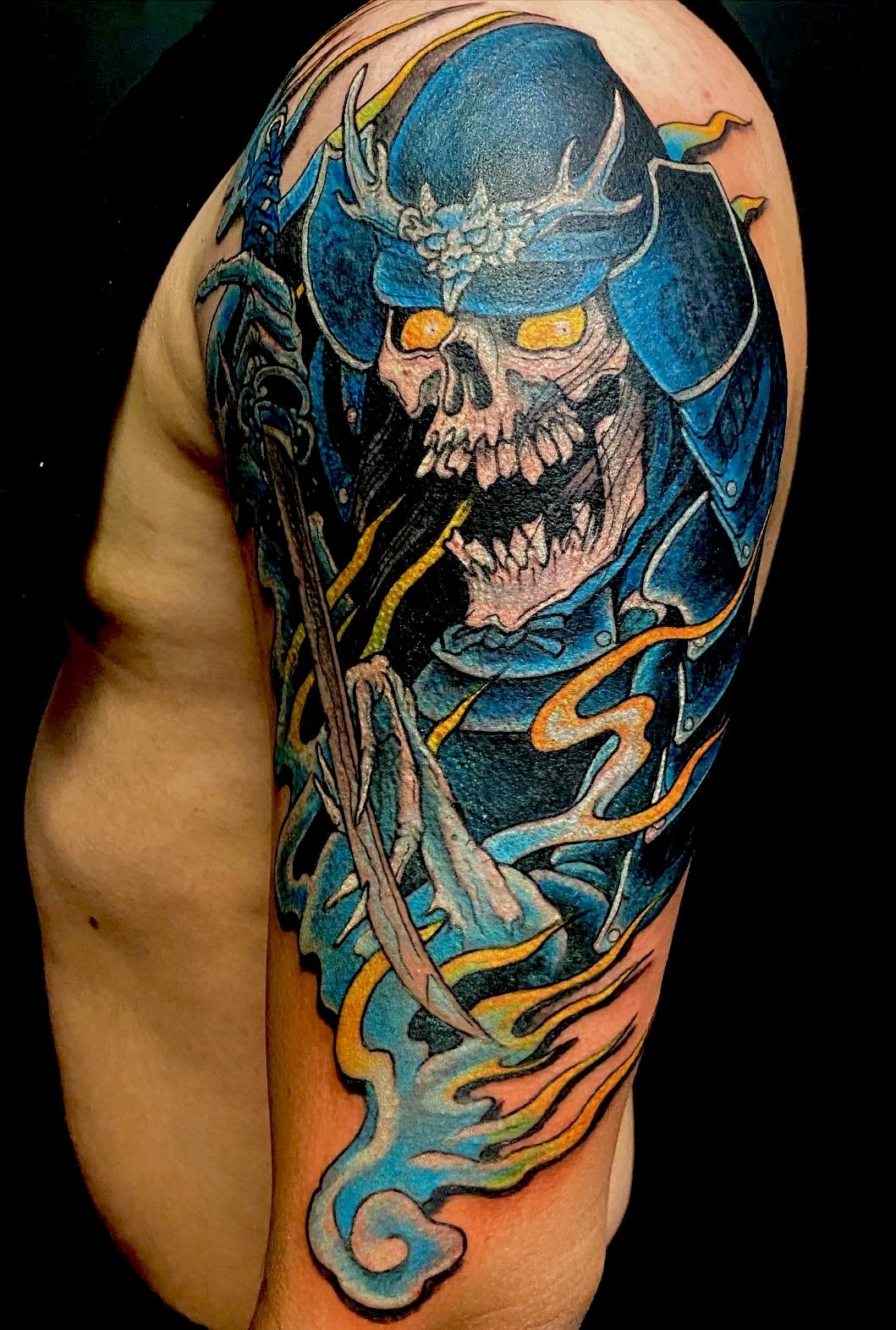 Colorful tattoo of a skeleton with yellow eyes wearing a helmet and armor, with flames and swirling patterns on the upper arm.