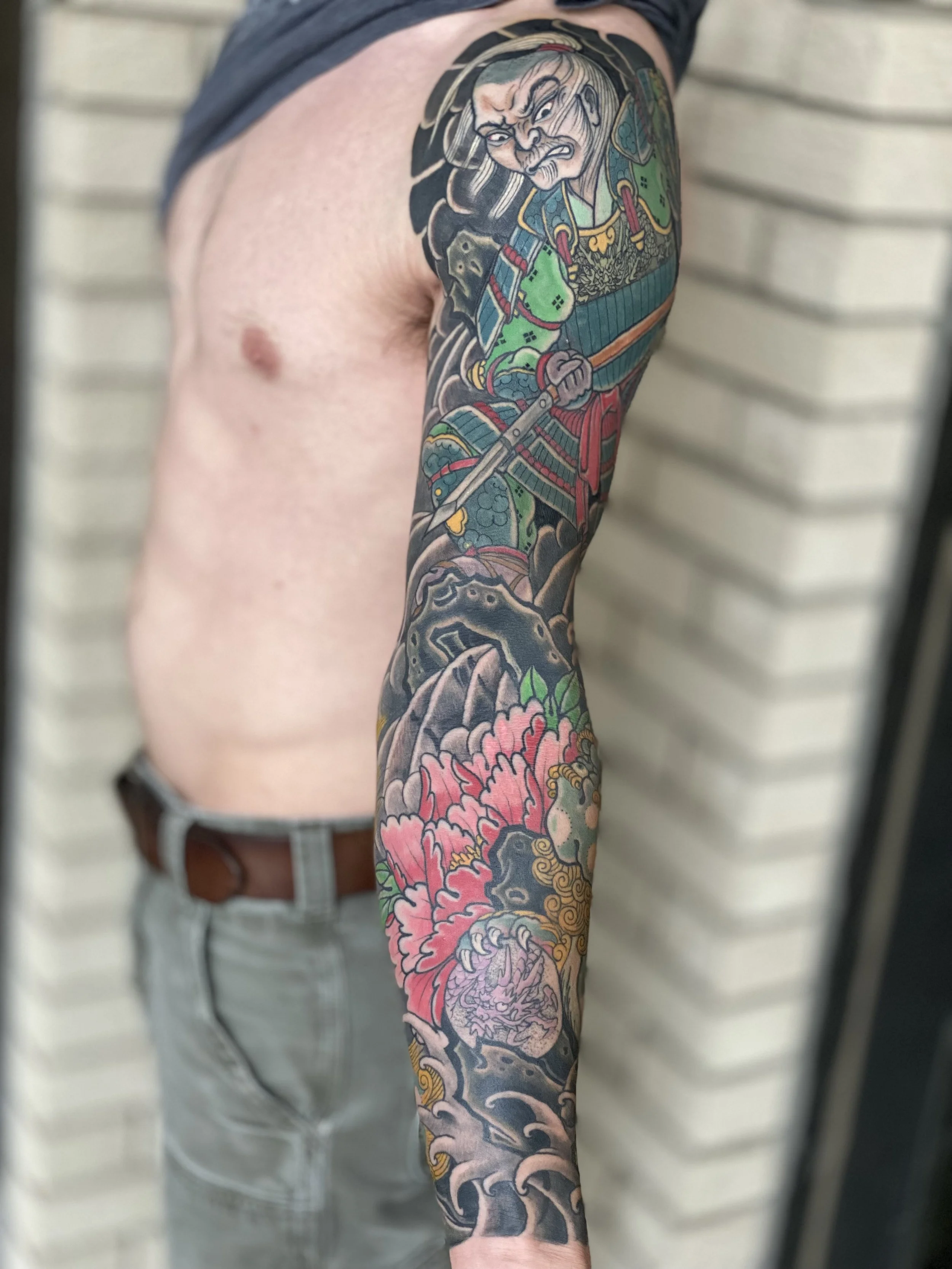 A person's arm with a colorful Japanese-style tattoo featuring a warrior in traditional armor holding a sword, surrounded by pink and green floral elements and waves.