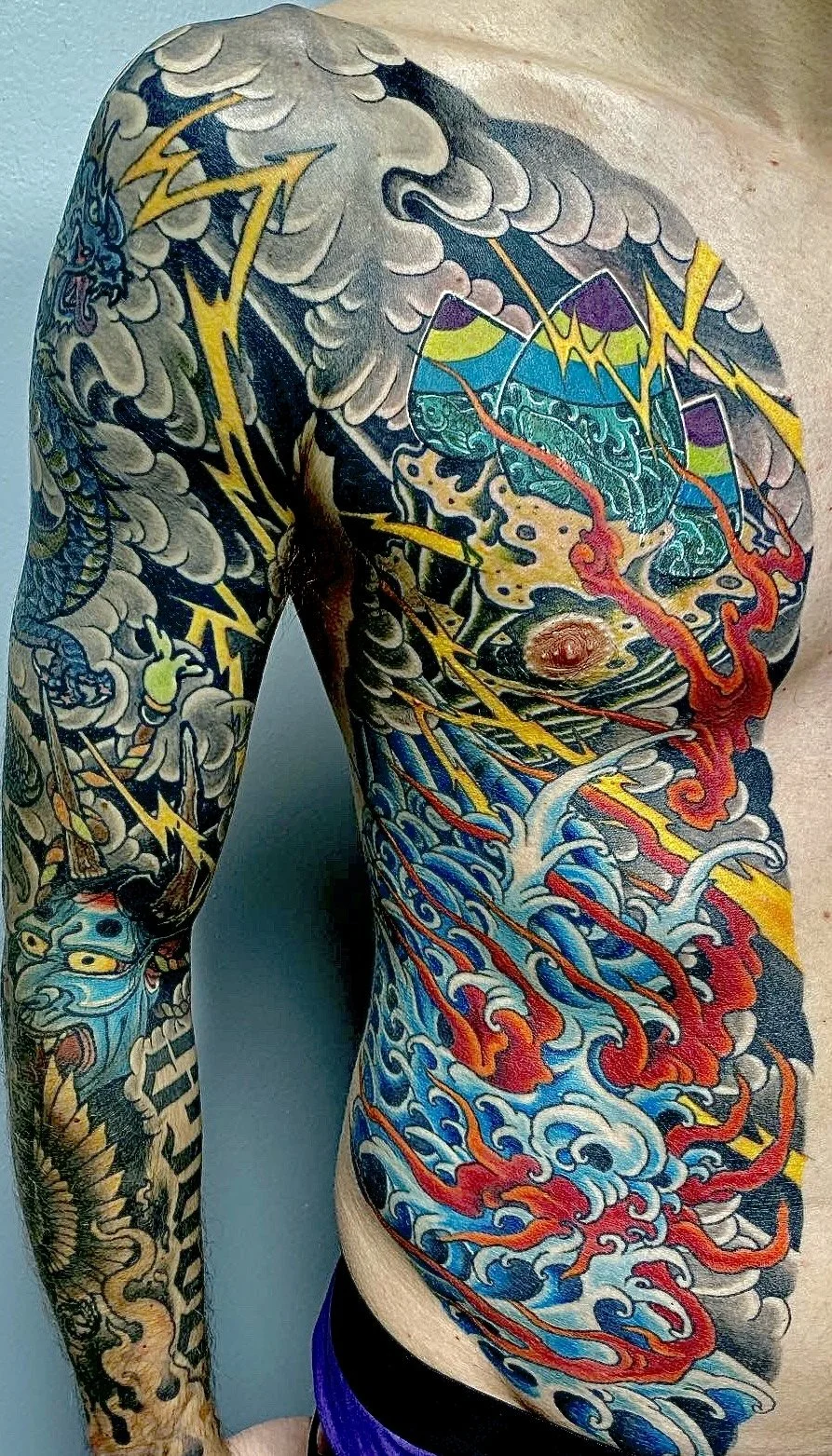 Colorful full sleeve tattoo featuring a dragon, storm clouds, lightning bolts, mountains, a rising sun, ocean waves with red and blue water, and abstract patterns.