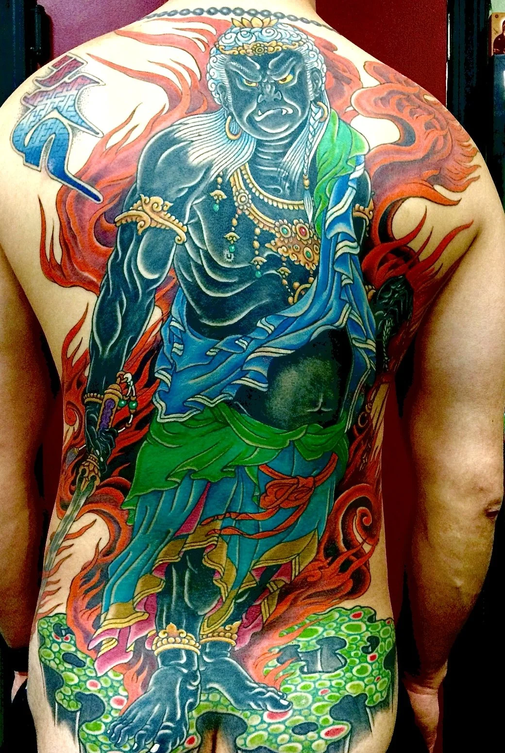 A colorful tattoo of a fierce, mythical warrior figure with dark blue skin, glowing yellow eyes, and long white hair, surrounded by red and orange flames, with green foliage at the bottom.