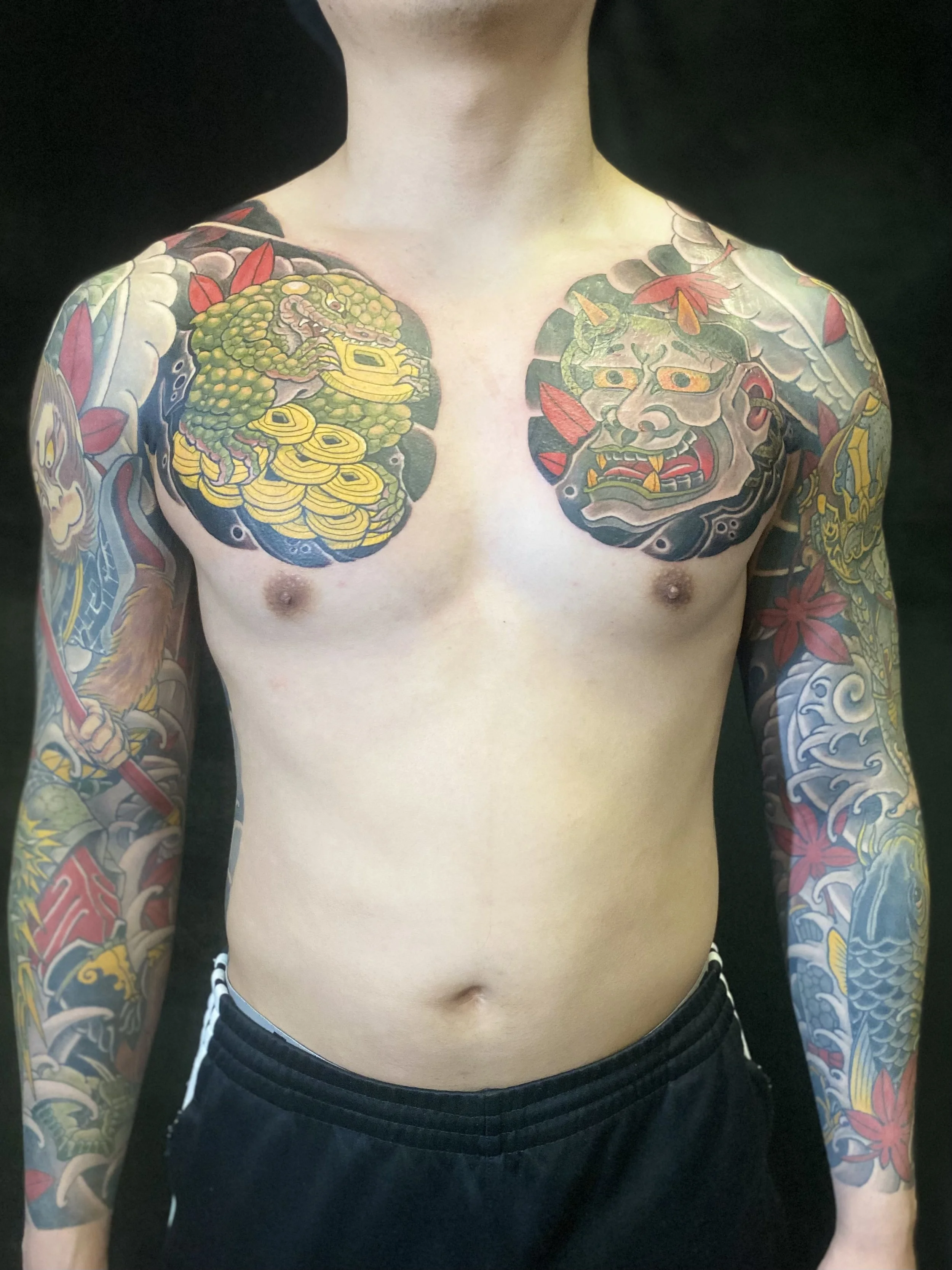 A shirtless person with extensive traditional Japanese style tattoos on both shoulders and arms, featuring a dragon, a demon mask, and colorful floral elements.