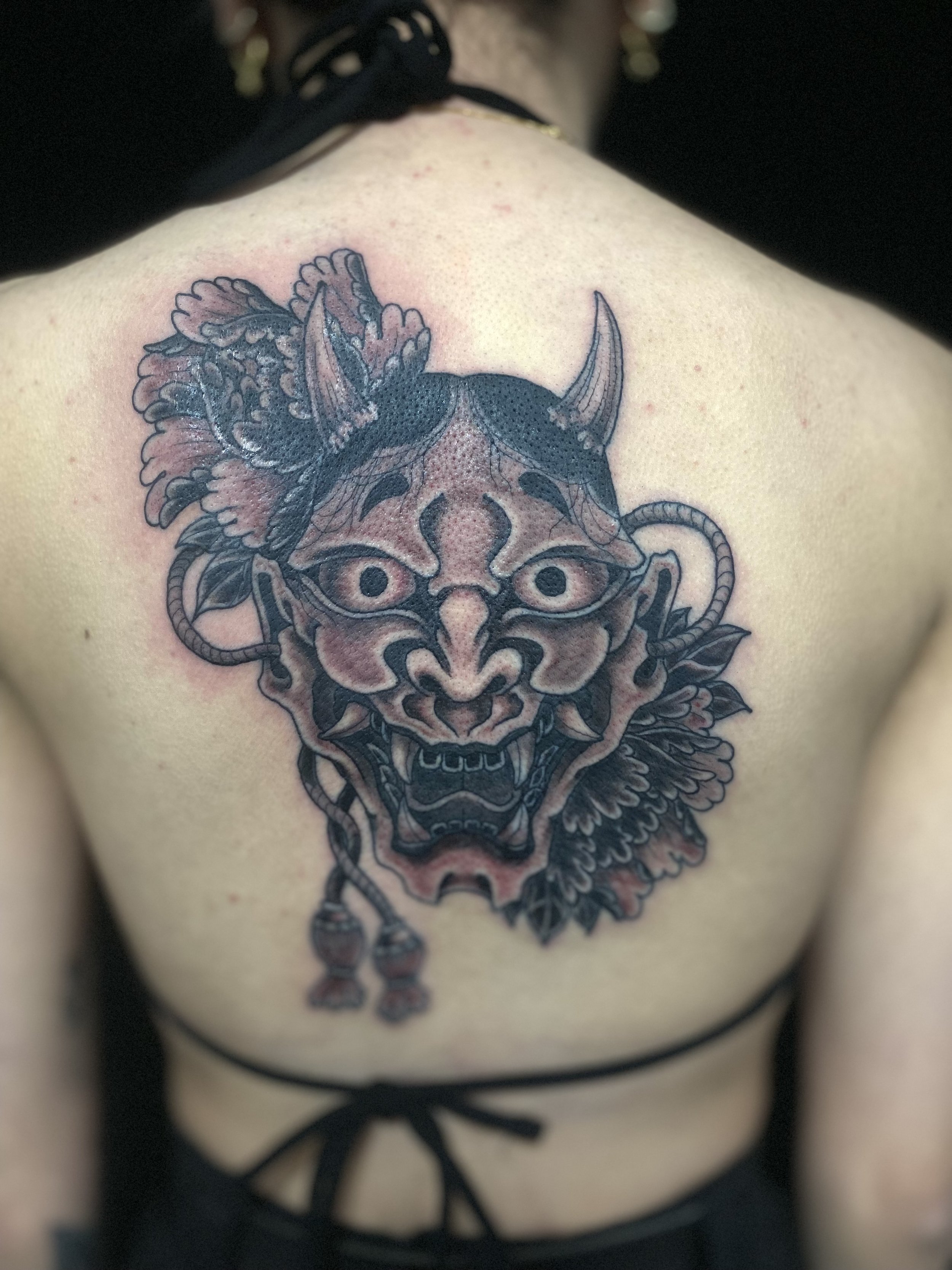 Tattoo of a fierce demon mask with horns, surrounded by detailed floral and feather elements, on a person's back.