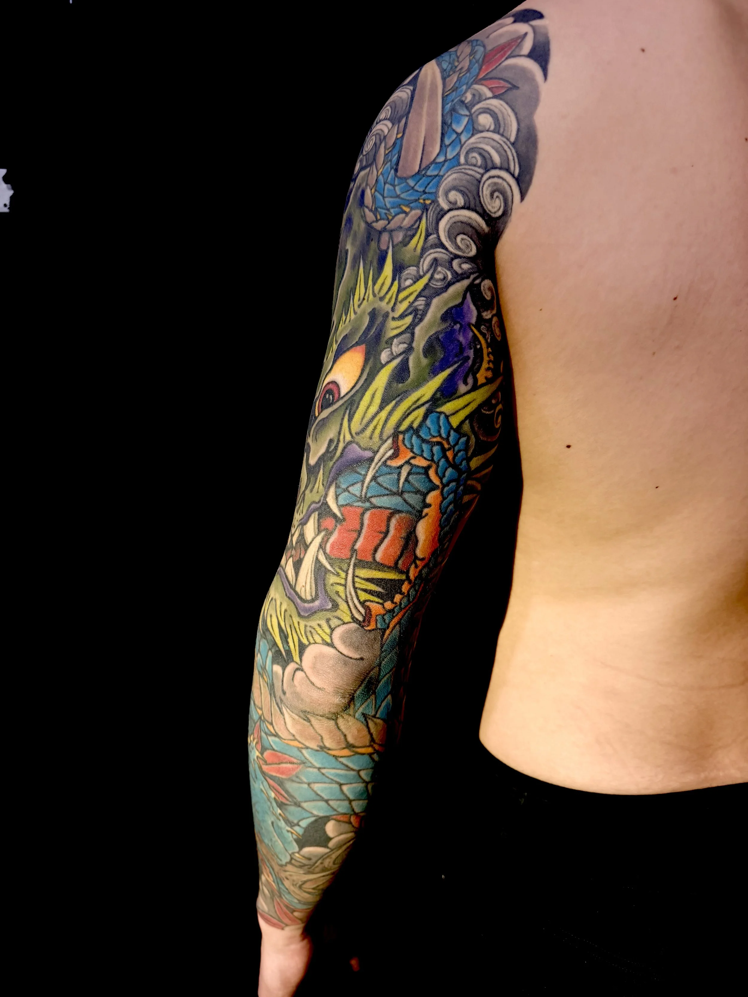 A person's upper arm sleeved with colorful traditional Japanese tattoo artwork, including a dragon and waves, against a black background.