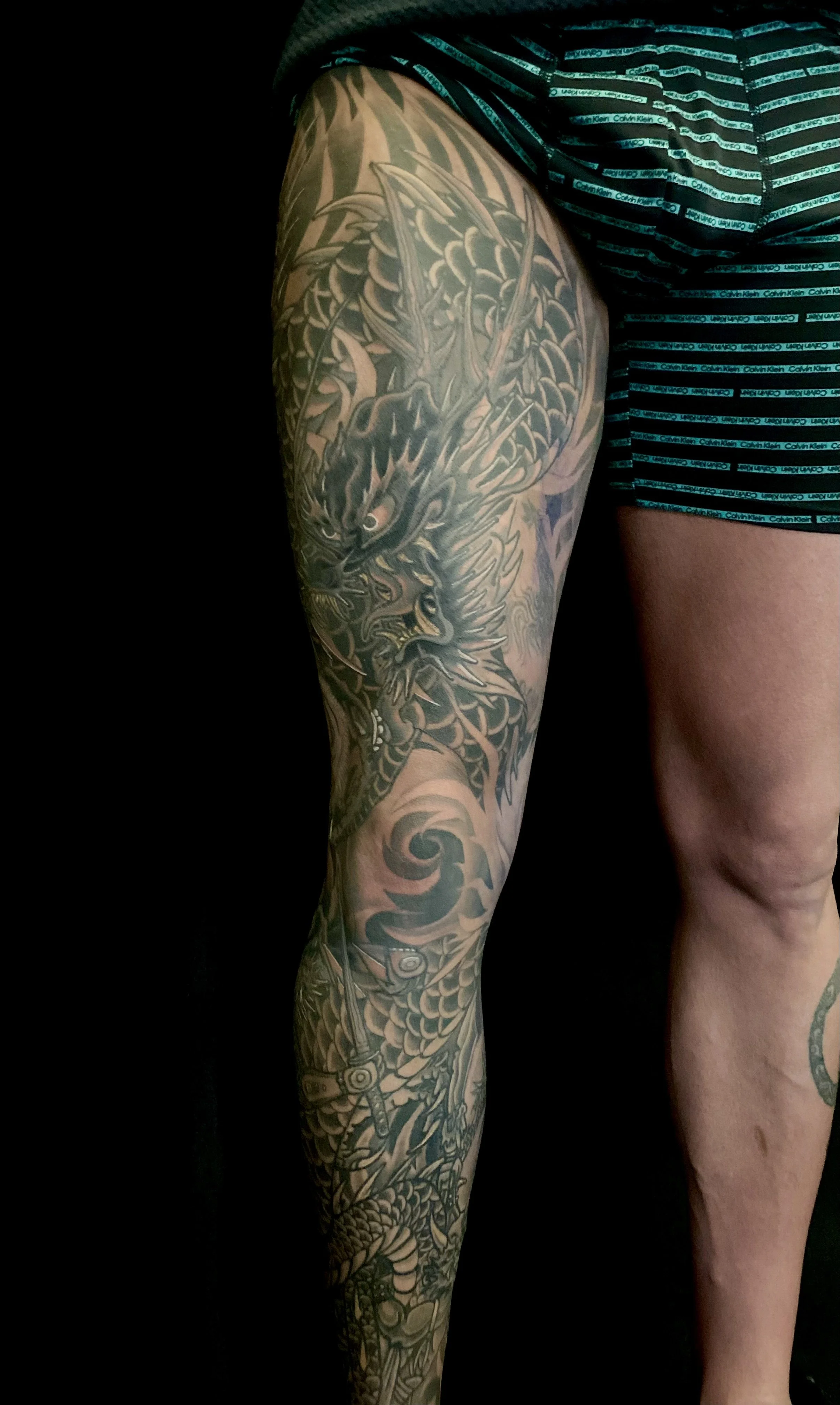 Close-up of a person's tattooed right thigh and hip, featuring a detailed dragon design in black and gray ink, with scales, claws, and a fierce expression.