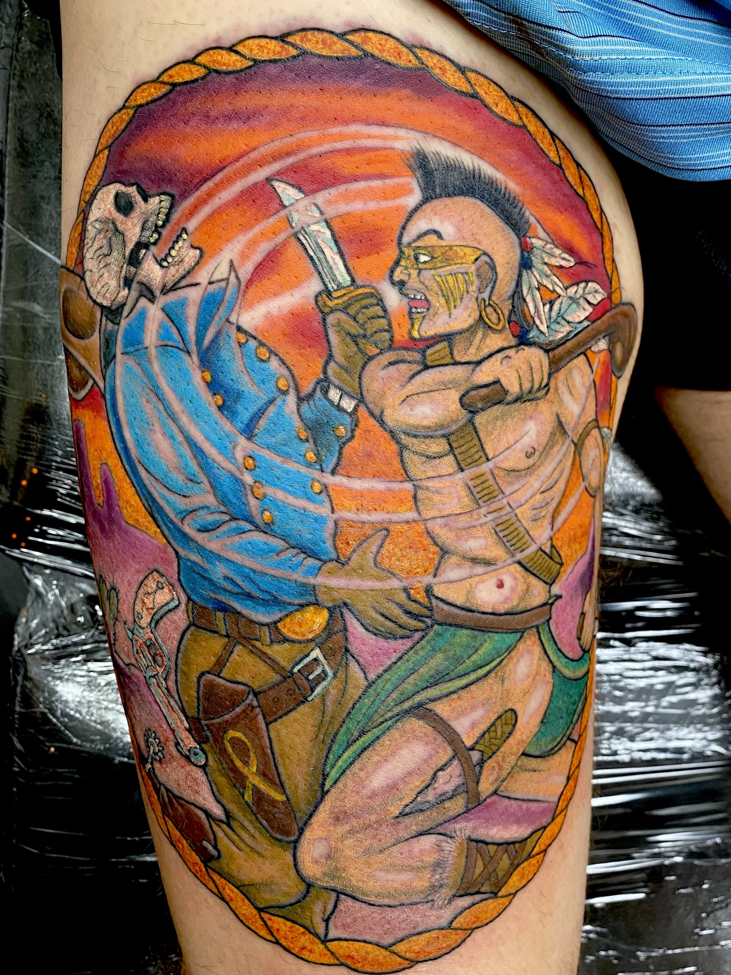 A colorful tattoo depicting a Native American warrior in traditional attire, holding a knife and facing a skull with a bone in its mouth. The scene is framed by a braided border and features vibrant orange, blue, and green hues.