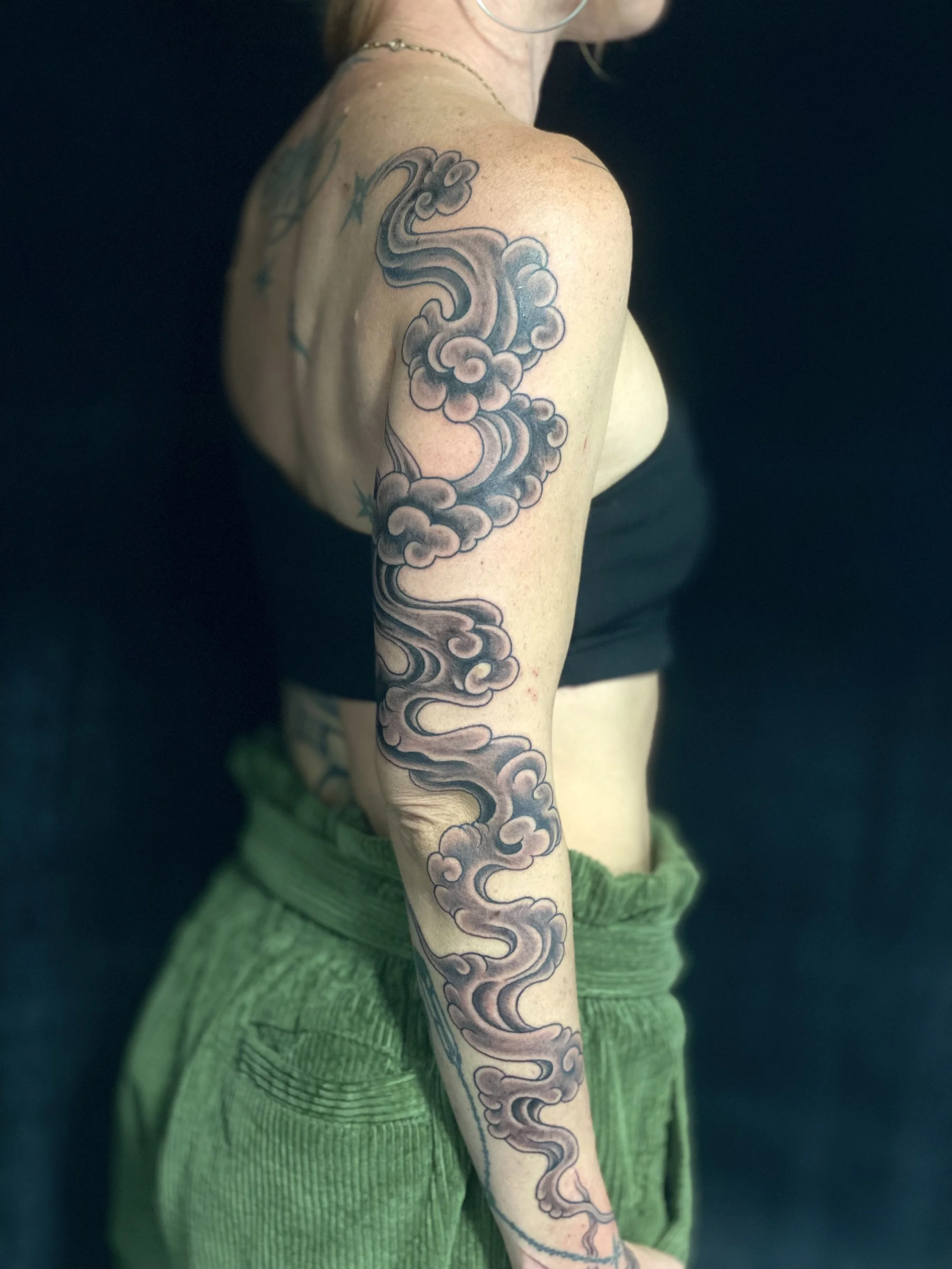 A woman with a large black and gray cloud and smoke tattoo on her left arm, wearing a black tube top and green pants, captured from her shoulder to her waist against a dark background.