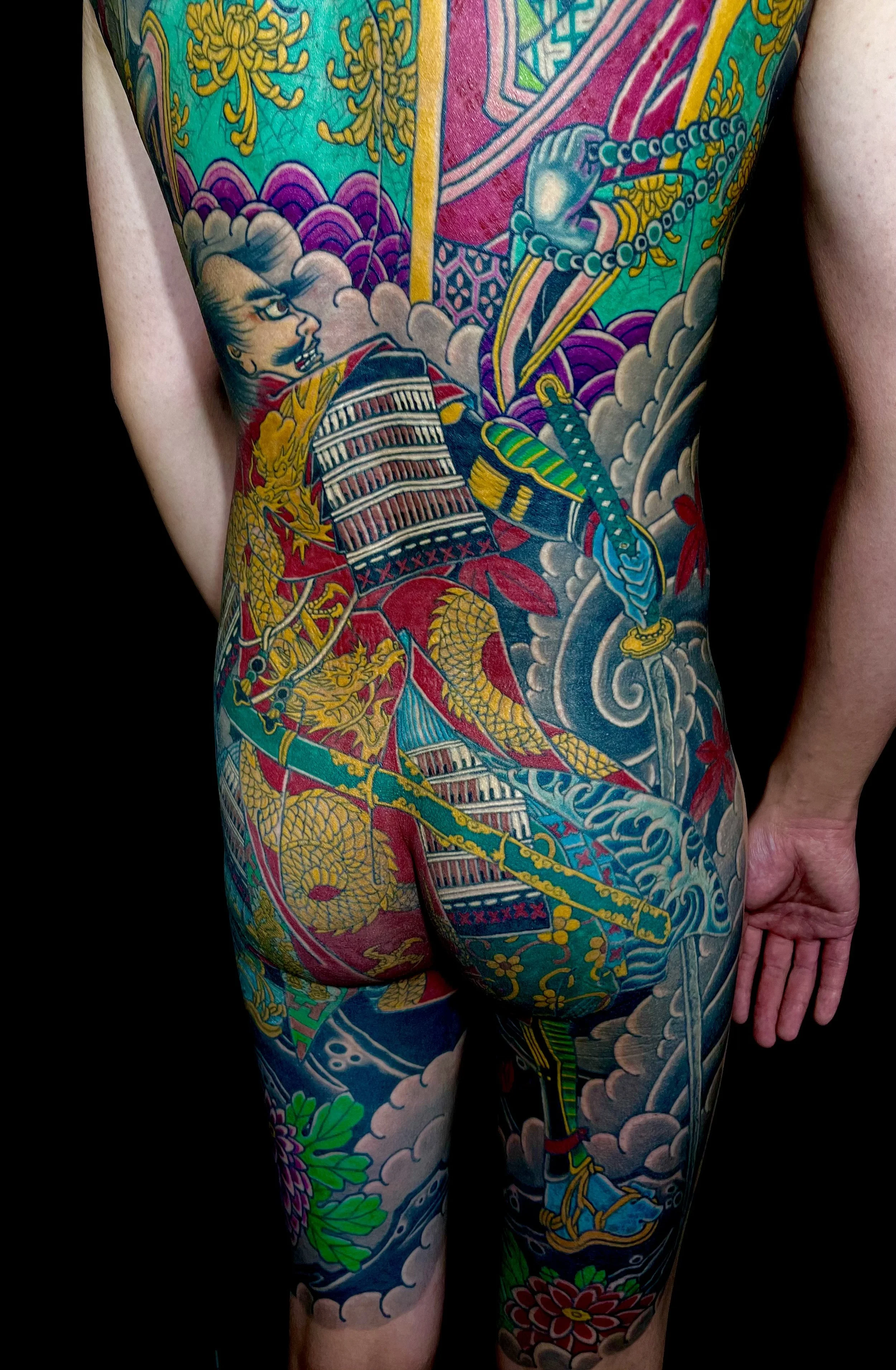 A person with colorful traditional Japanese-style tattoos covering their back, arms, and legs, featuring imagery of a woman, dragons, swords, flowers, and clouds.