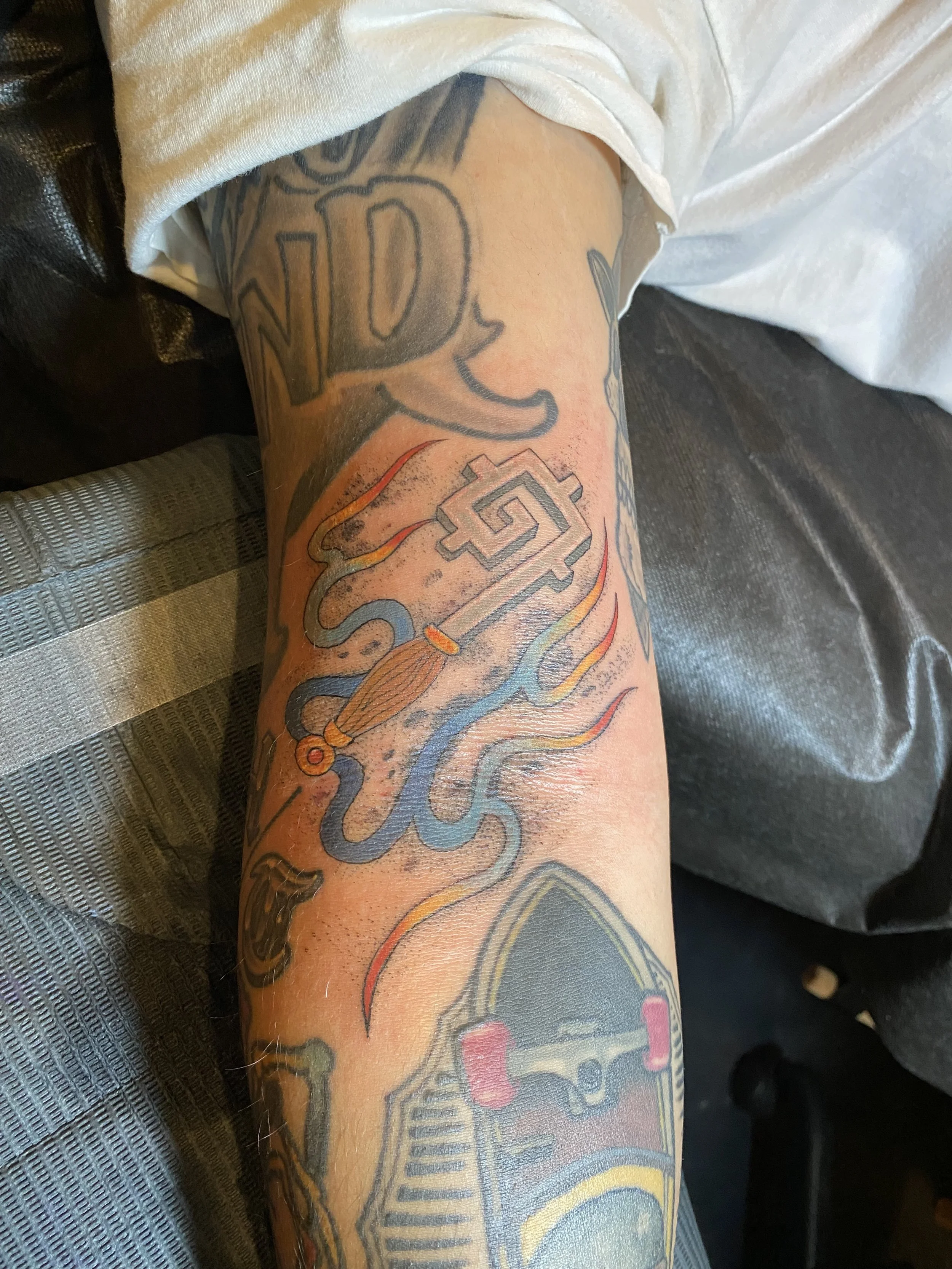 A person's arm with multiple colorful tattoos, including a skateboard, flames, a snake, and various abstract designs.