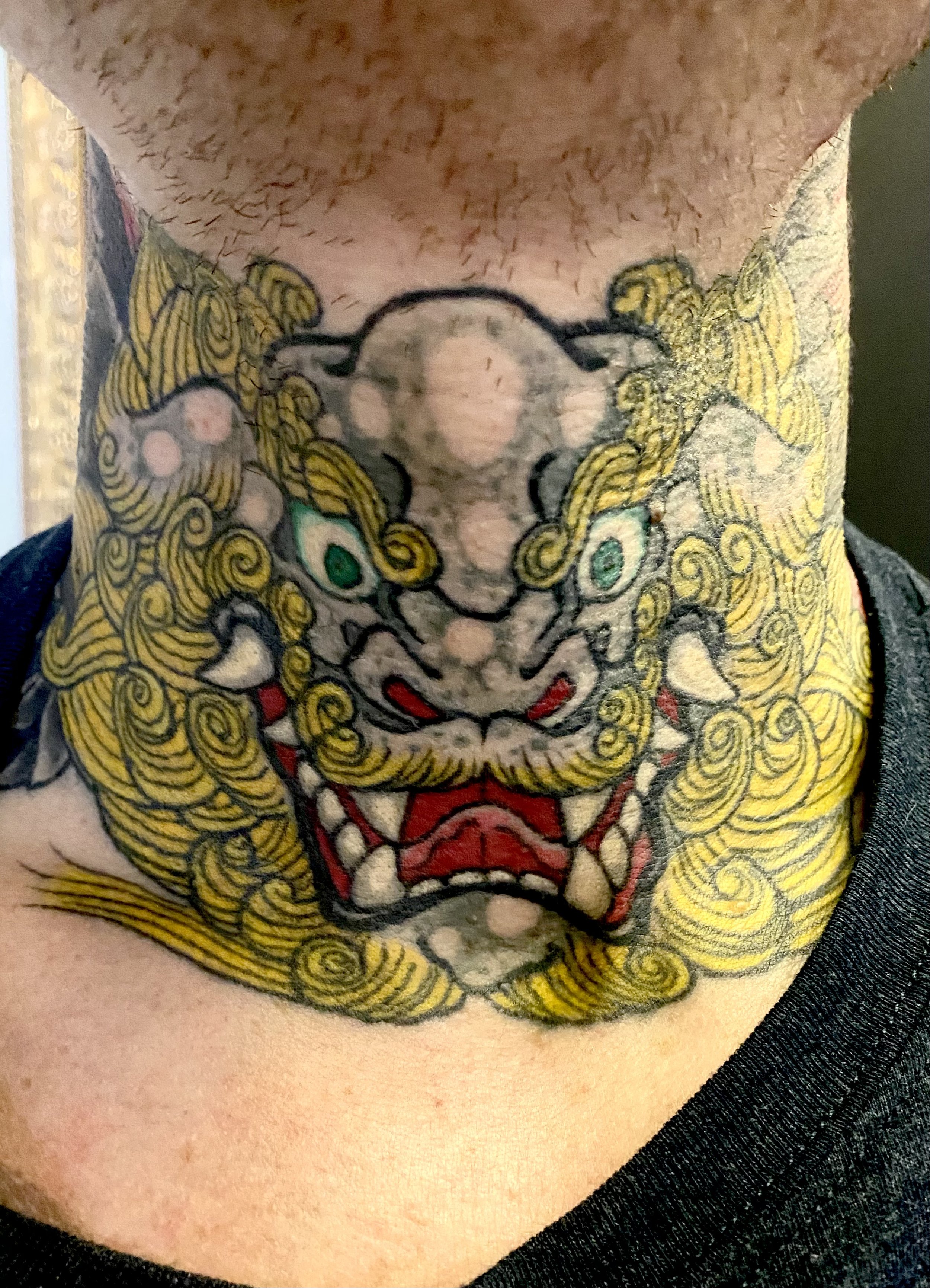 Colorful tattoo of a fierce lion's face with intricate yellow mane, green eyes, and red tongue on a person's neck.
