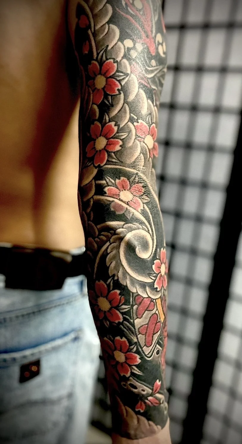 Close-up of a person's arm with a colorful floral tattoo sleeve, featuring pink and yellow flowers and green leaves, against a blurred background of black and white grid-like structure.