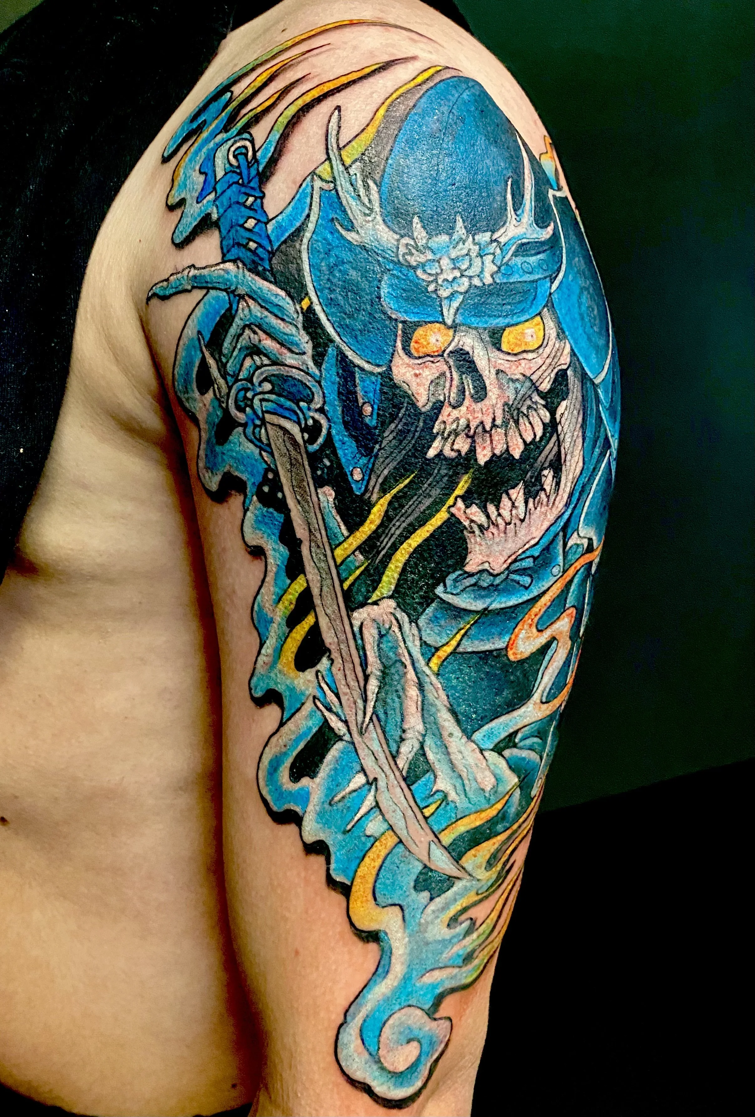 Colorful tattoo of a skull wearing a demon mask and a helmet, wielding a sword with swirling blue and yellow flames, covering a person's upper arm and shoulder.