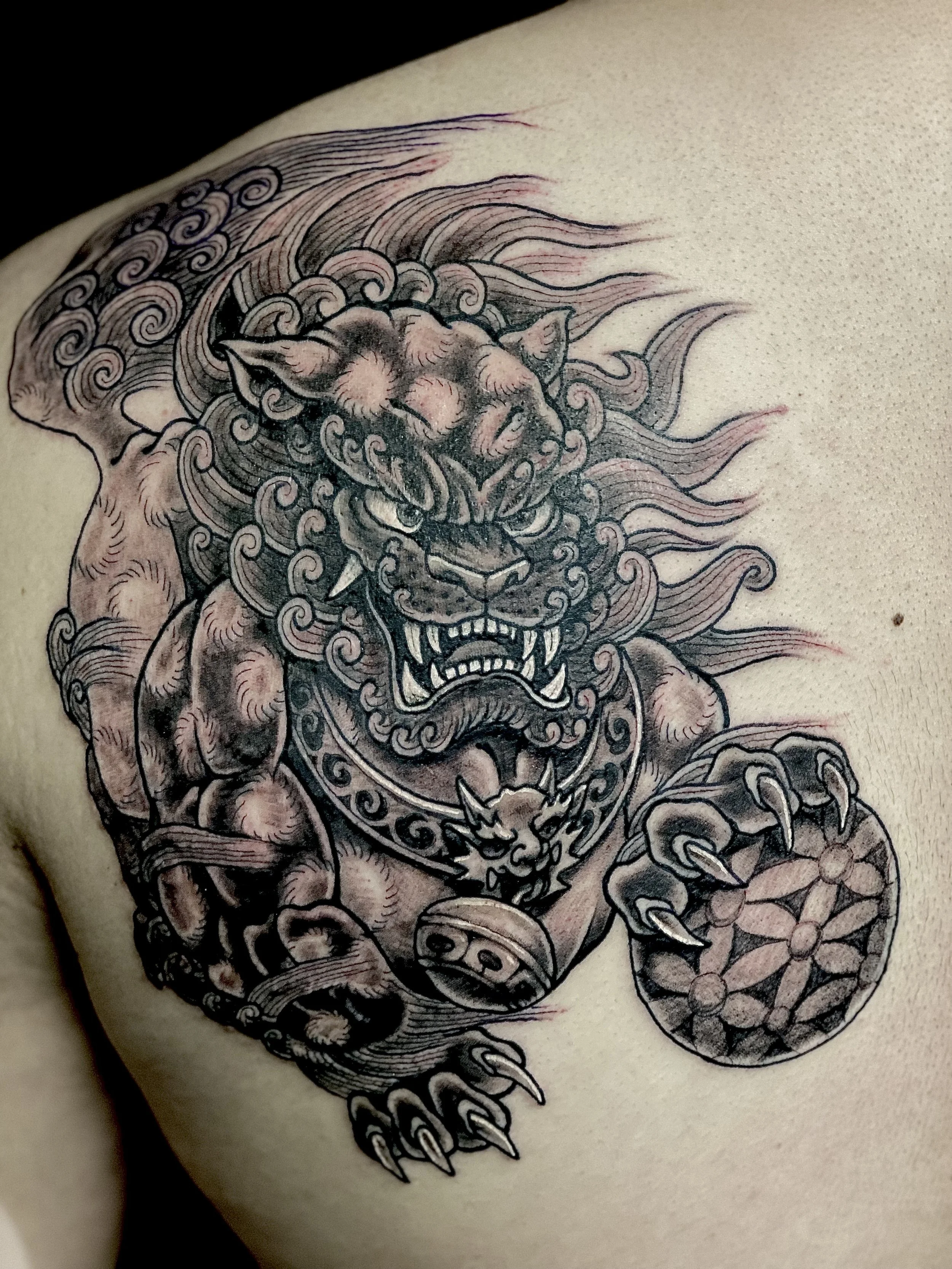 Tattoo of a fierce lion with a muscular body, large claws, and a circular shield with a geometric pattern.