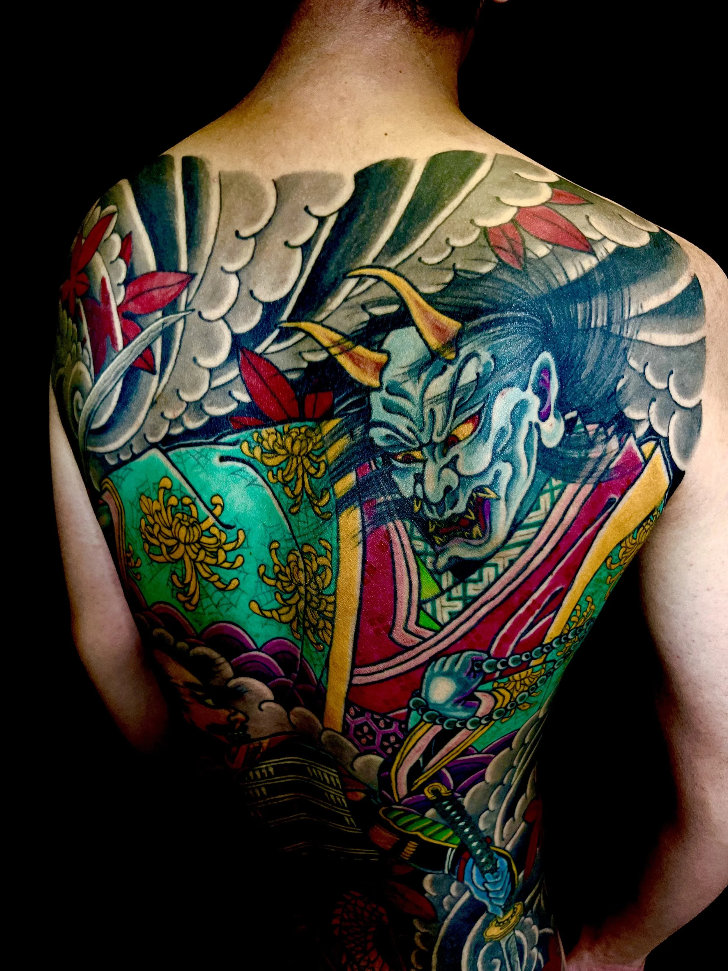 Colorful traditional Japanese-style tattoo featuring a fierce demon or Oni with a sword, surrounded by clouds and flames.