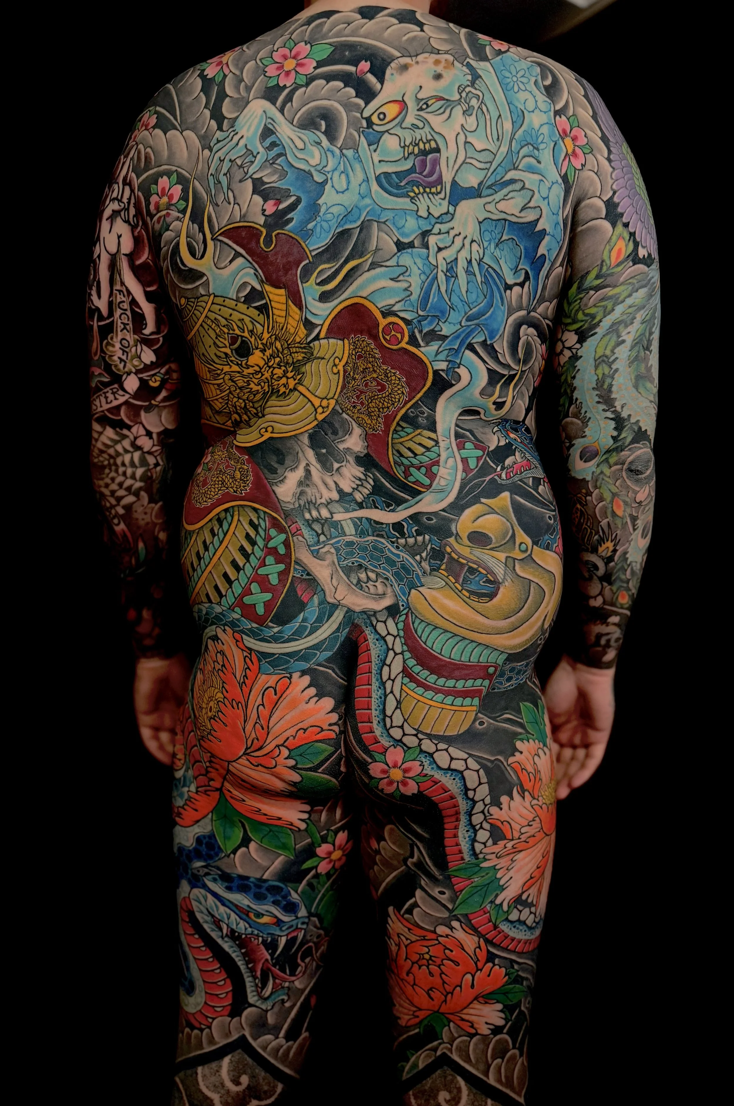 Full-back view of a tattoo sleeve illustration of a fierce blue dragon, a fierce yellow tiger, pink cherry blossoms, and various traditional Japanese motifs against a dark background.