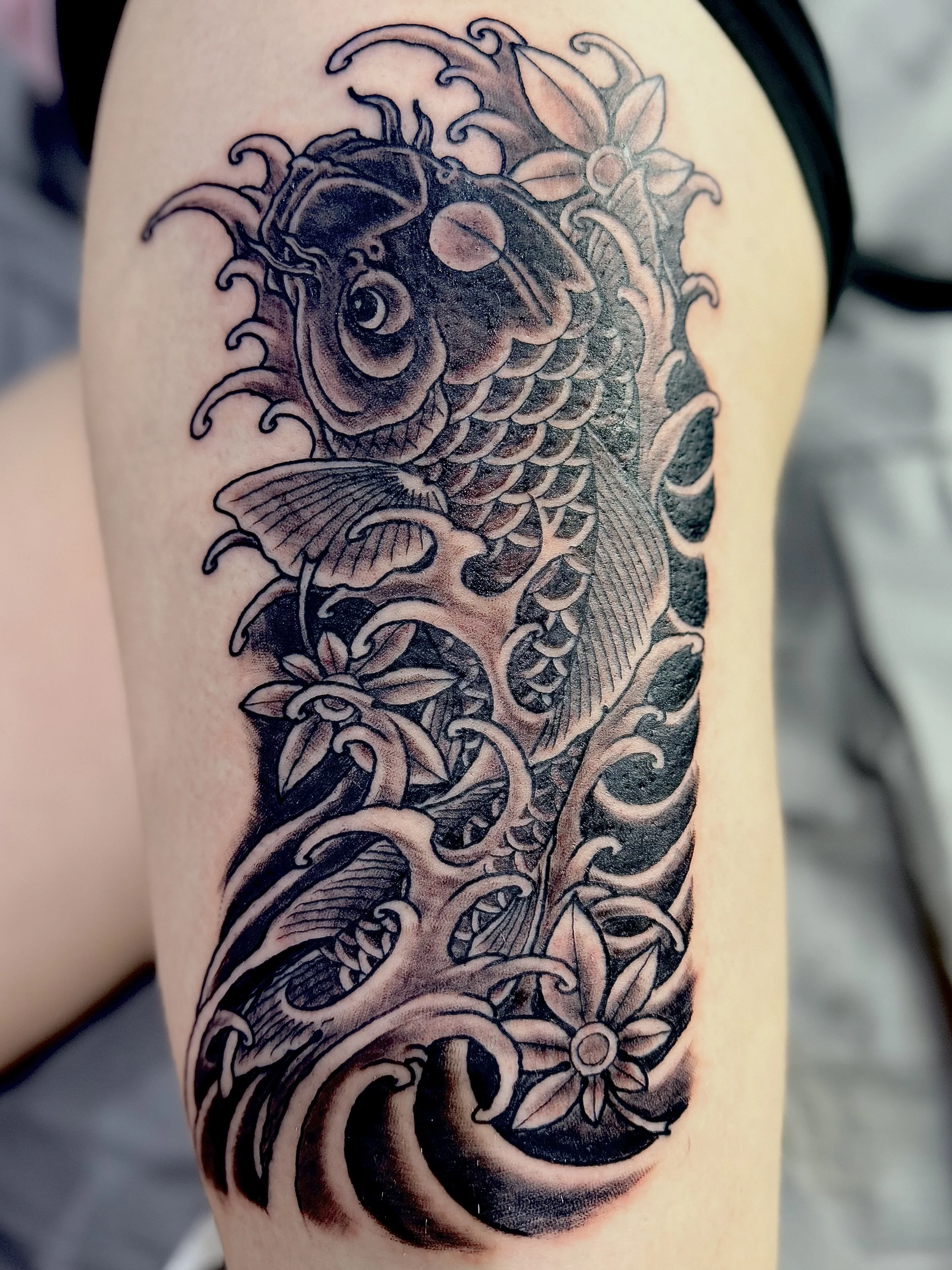 Black and gray tattoo of a koi fish with flowers on a person's arm.