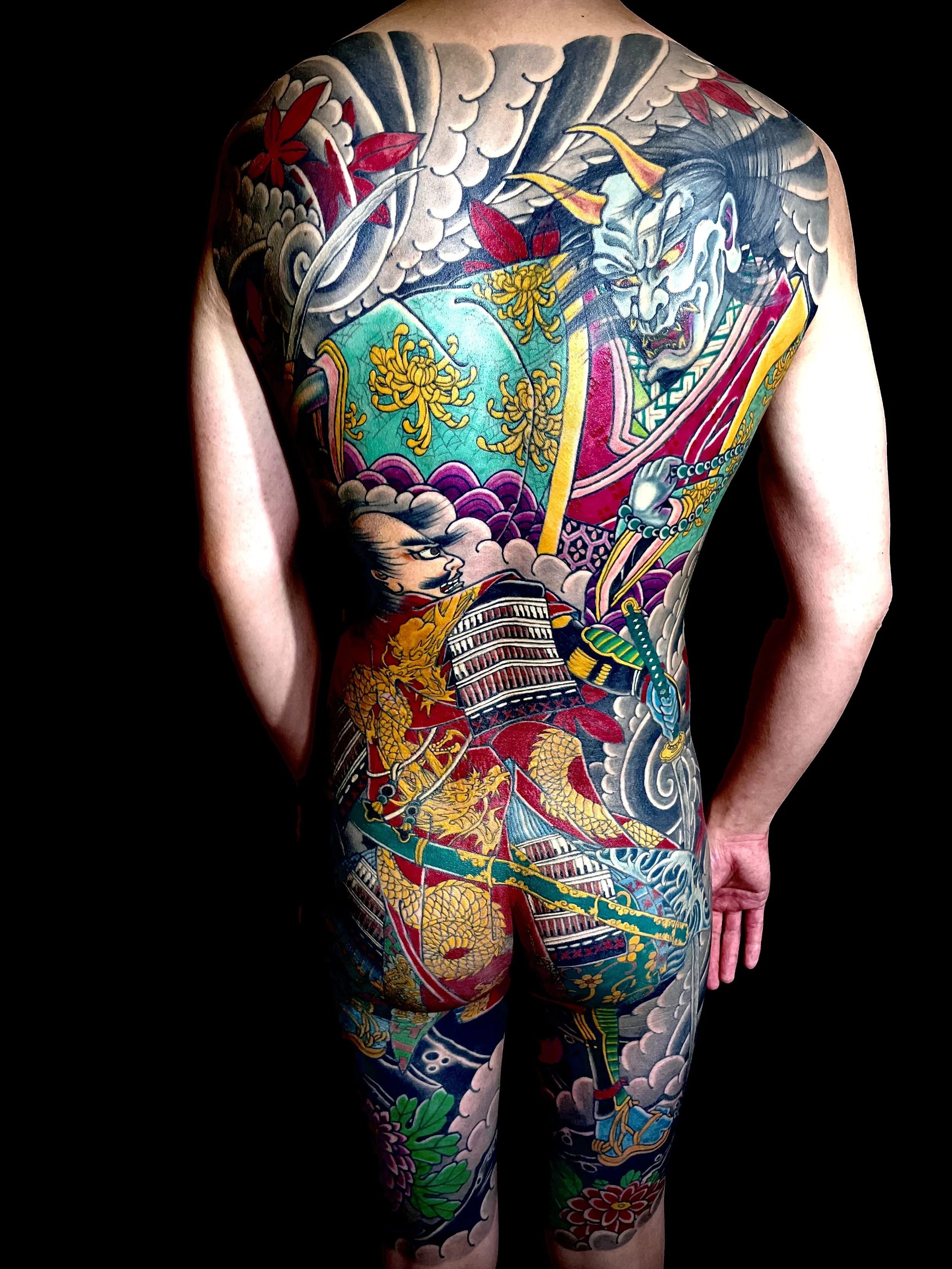 Colorful traditional Japanese tattoo covering the person's back, shoulders, and legs depicting a fierce samurai warrior with a demon mask, surrounded by clouds, flowers, and Japanese motifs.