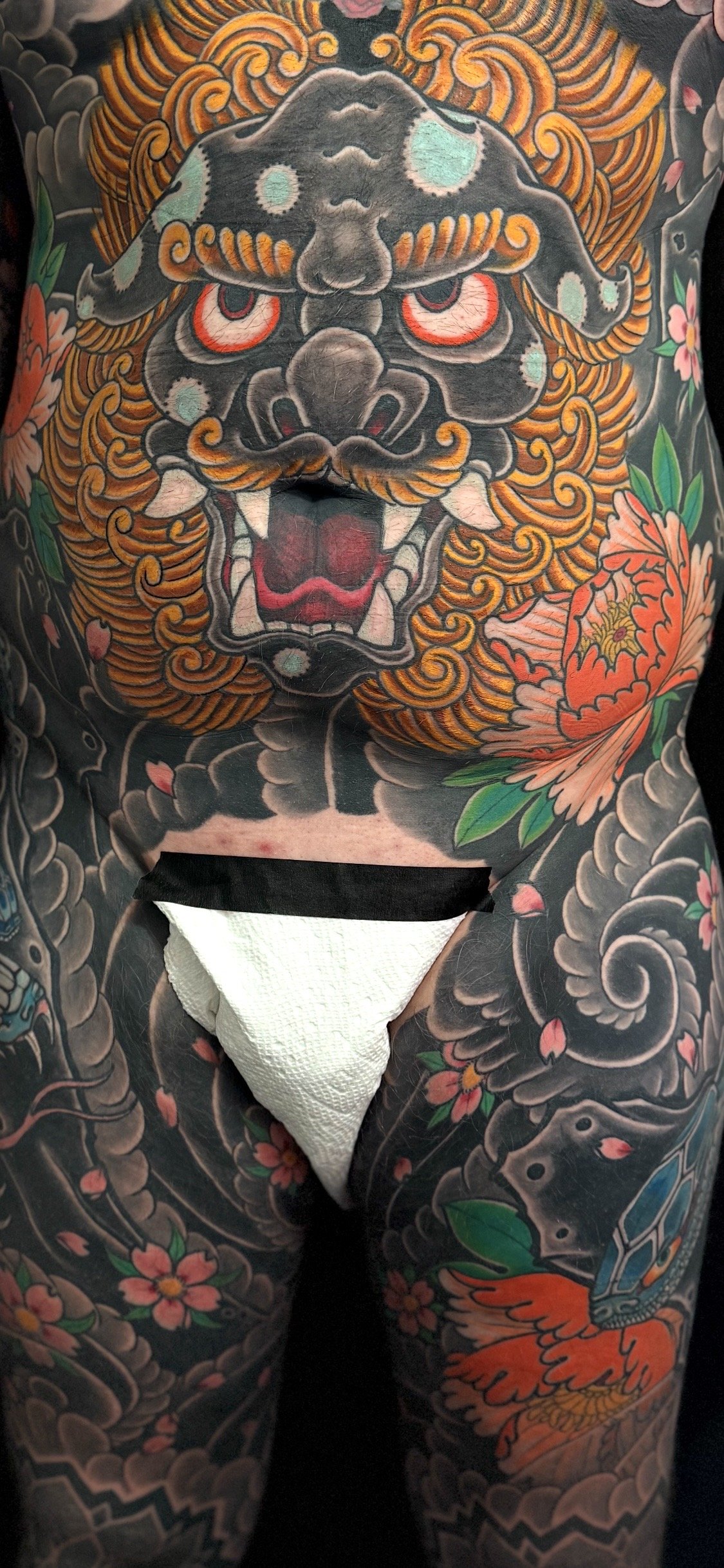 A person with extensive traditional Japanese-style tattoo art covering the chest and arms, featuring a fierce, colorful lion or dragon face with red eyes, an open mouth showing teeth, surrounded by floral and cloud motifs.