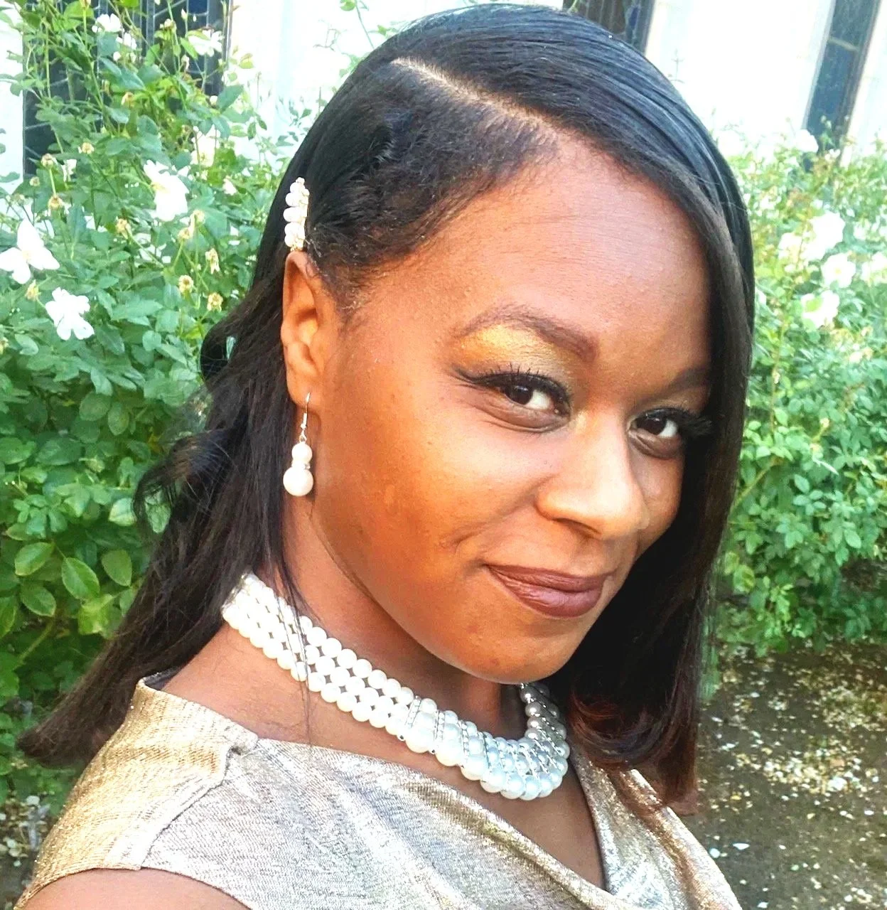 Woman wearing pearl necklace and gold dress with garden background.