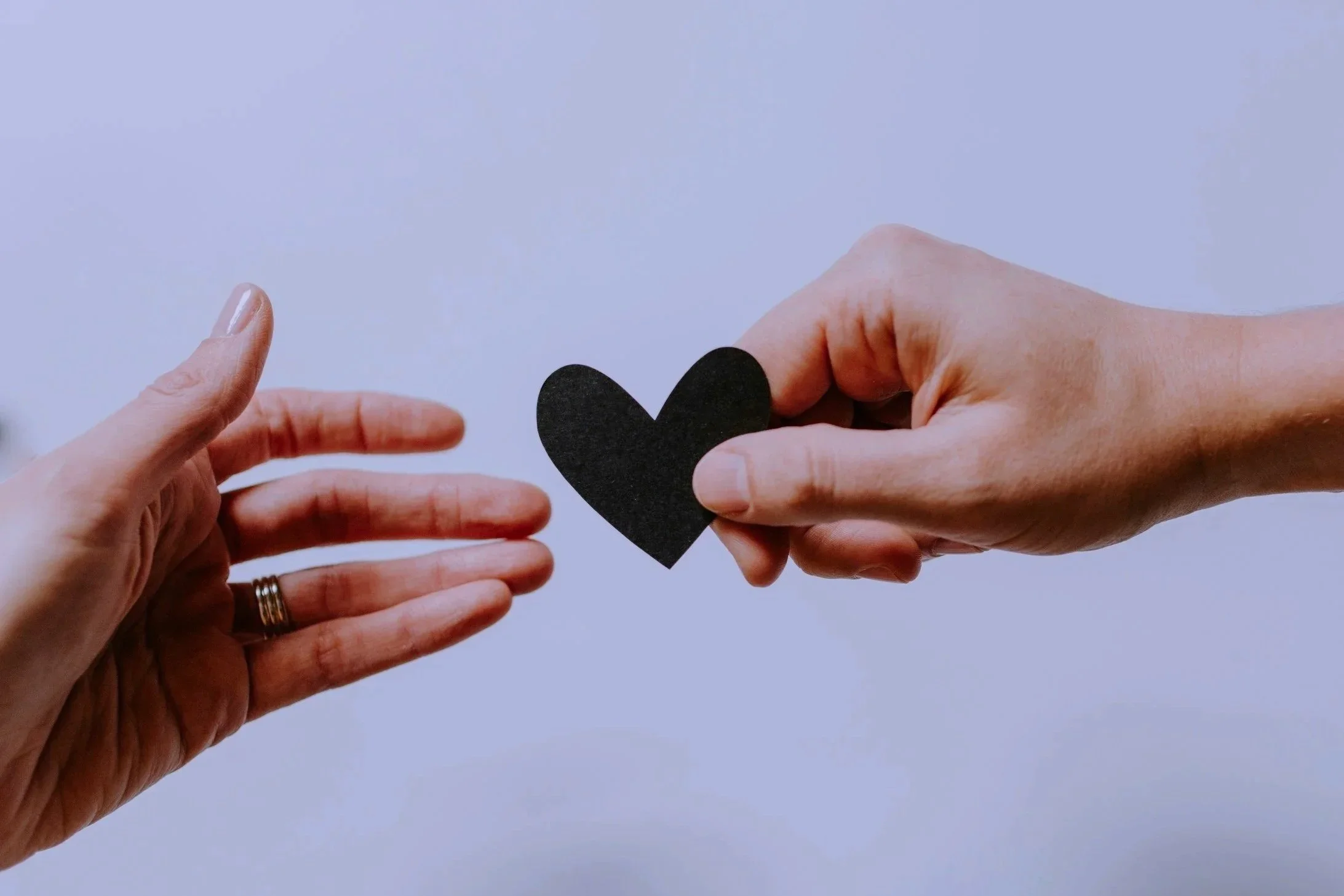 Two hands, one with a ring, exchanging a black paper heart against a plain light background.