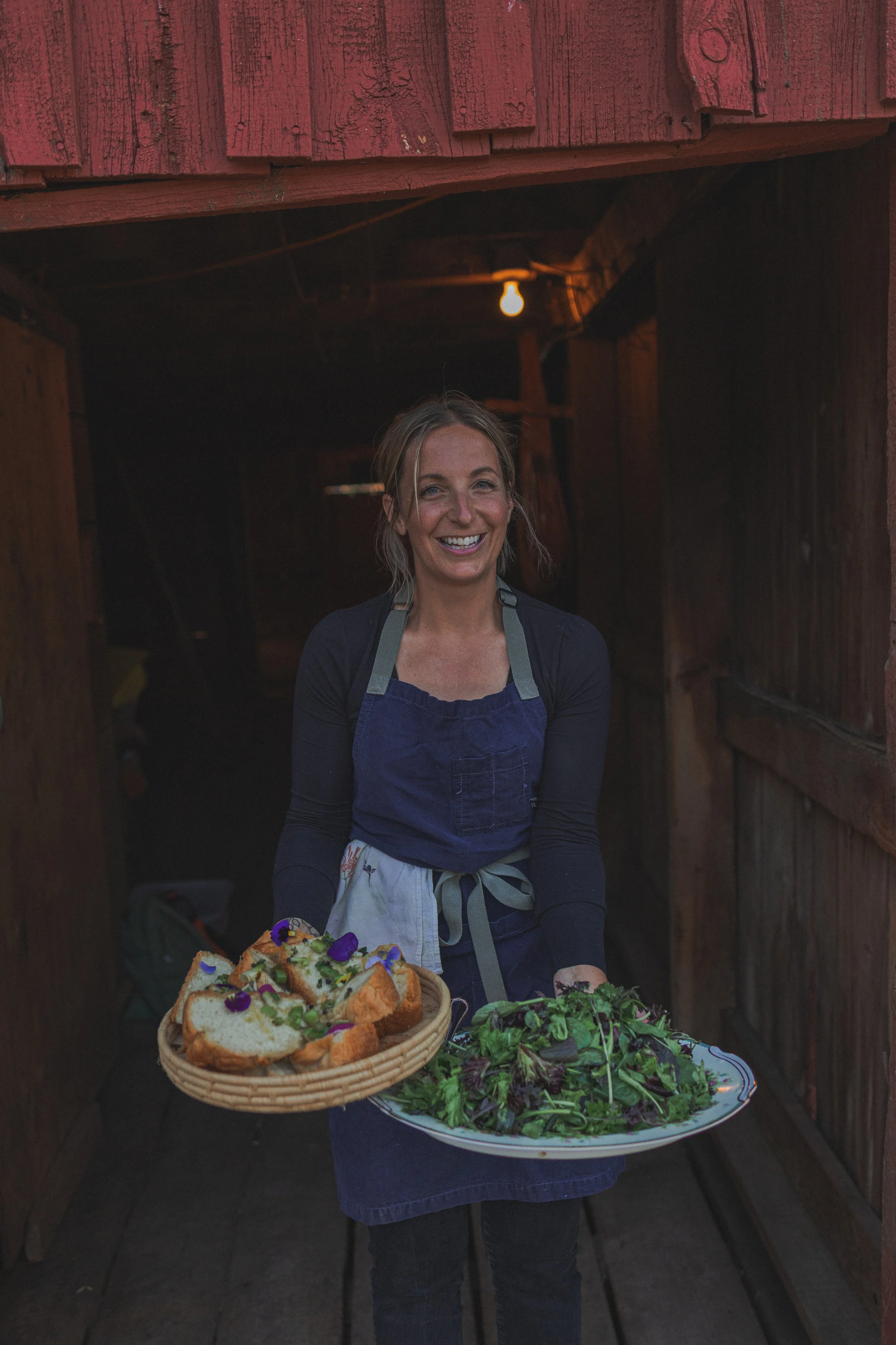 A woman smiling and carrying plates of food, including slices of bread with toppings and a large salad, in a rustic wooden setting.